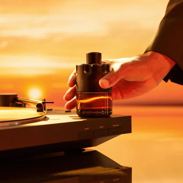 Frasco âmbar escuro do perfume The Most Wanted da Azzaro sendo segurado por mão masculina sobre toca-discos, com pôr do sol ao fundo evocando calor e intensidade.