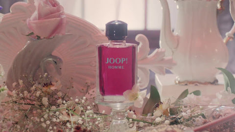 Frasco do perfume Joop! Homme em destaque sobre uma superfície espelhada decorada com flores delicadas e elementos clássicos em porcelana branca. A composição suave e romântica contrasta com o tom vibrante do frasco rosa, evocando elegância e charme.