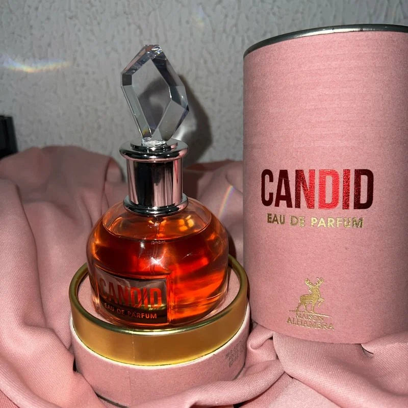 Frasco do perfume Candid Eau de Parfum da marca Maison Alhambra, com líquido âmbar e tampa transparente em formato geométrico. Está apoiado em sua caixa cilíndrica rosa com detalhes dourados, sobre um tecido rosa claro.