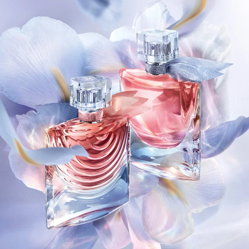 Dois frascos do perfume La Vie Est Belle da Lancôme entre pétalas roxas e azuis translúcidas, criando visual etéreo e feminino, com frascos curvos e delicados.