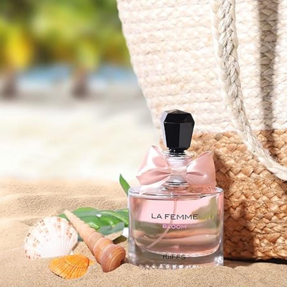 Frasco do perfume La Femme Bloom da marca Riiffs, com líquido rosa claro, tampa preta e laço rosa. O frasco está apoiado na areia de uma praia, ao lado de conchas, folhas e uma bolsa de palha, com fundo desfocado mostrando o mar e coqueiros ao longe.