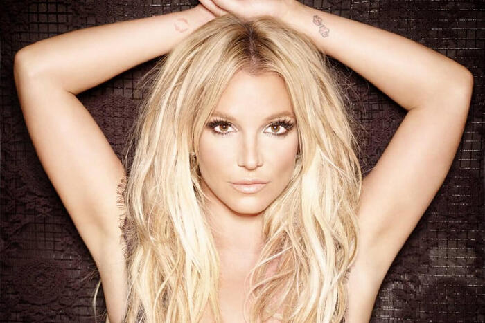 Britney Spears posando com os braços levantados acima da cabeça. Ela está em frente a um fundo escuro com textura rendada, usando maquiagem suave com destaque para os olhos. A expressão é séria e confiante, com olhar direto para a câmera.