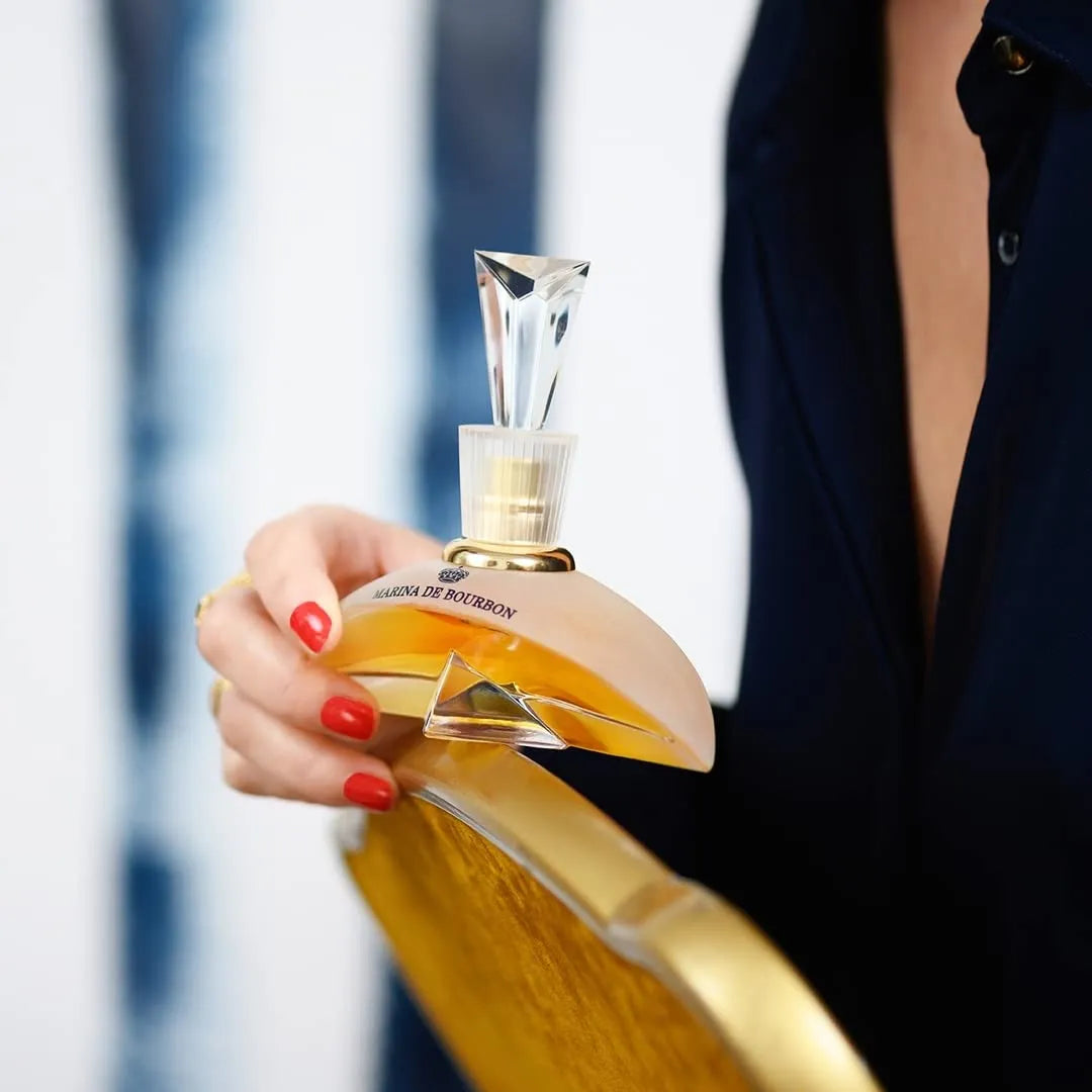 Frasco do perfume Marina de Bourbon Rouge Royal Elite com design dourado e tampa em formato de cristal, sendo segurado por uma mulher com unhas vermelhas e traje azul-marinho, em um cenário sofisticado e moderno.