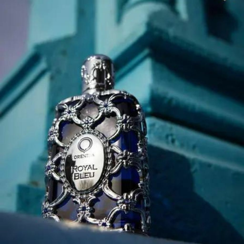 Frasco do perfume Royal Bleu da marca Orientica, com design luxuoso e detalhes prateados ornamentados envolvendo o vidro azul. A embalagem sofisticada contrasta com o fundo em tons de azul arquitetônico, transmitindo imponência e exclusividade.
