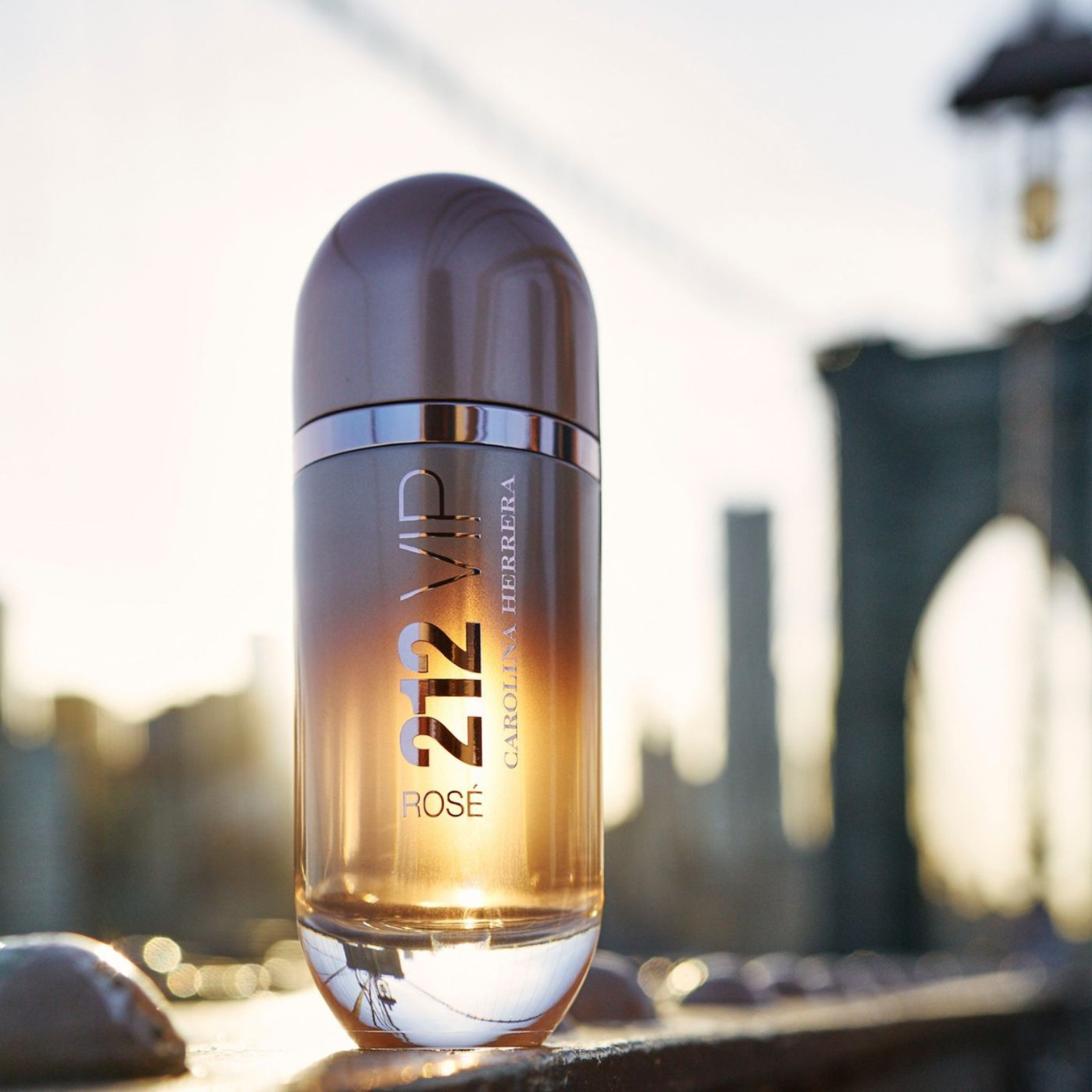 Perfume 212 VIP Rosé Carolina Herrera em primeiro plano com um fundo desfocado de uma paisagem urbana ao amanhecer, com tons dourados e a silhueta de uma ponte, sugerindo uma fragrância vibrante e elegante para a vida na cidade.