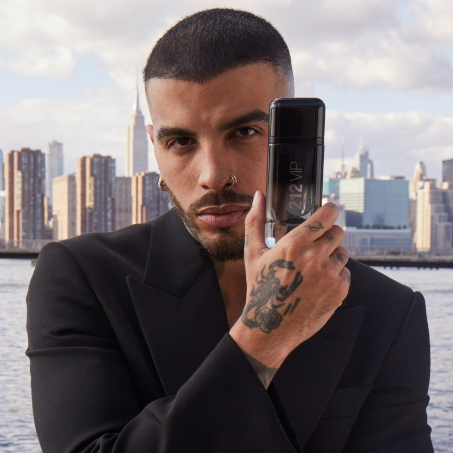 Homem elegante com tatuagem no braço segurando o perfume importado masculino 212 VIP Black Carolina Herrera em frente a um cenário urbano de Nova York, personificando o estilo sofisticado e confiante, disponível na Velyre.