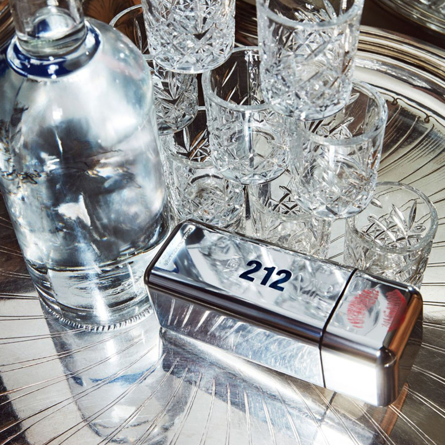Perfume 212 Vip Men Carolina Herrera posicionado em uma bandeja de metal prateado ao lado de diversos copos de cristal lapidados e uma garrafa, sugerindo uma atmosfera de celebração e elegância.