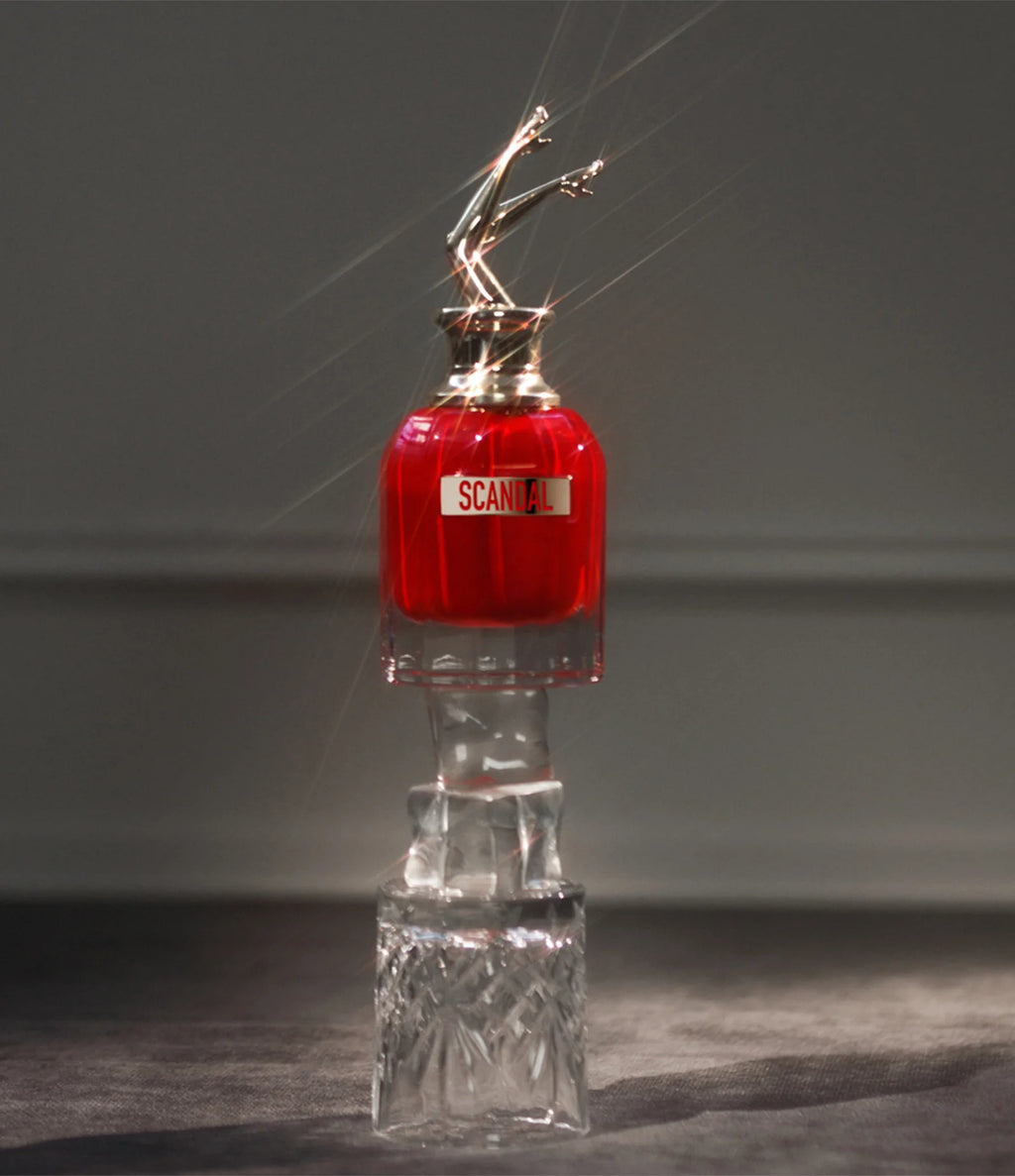 Frasco do Scandal Le Parfum iluminado por feixes de luz, posicionado sobre uma base de vidro esculpido, com destaque para o brilho intenso e sofisticado do frasco.