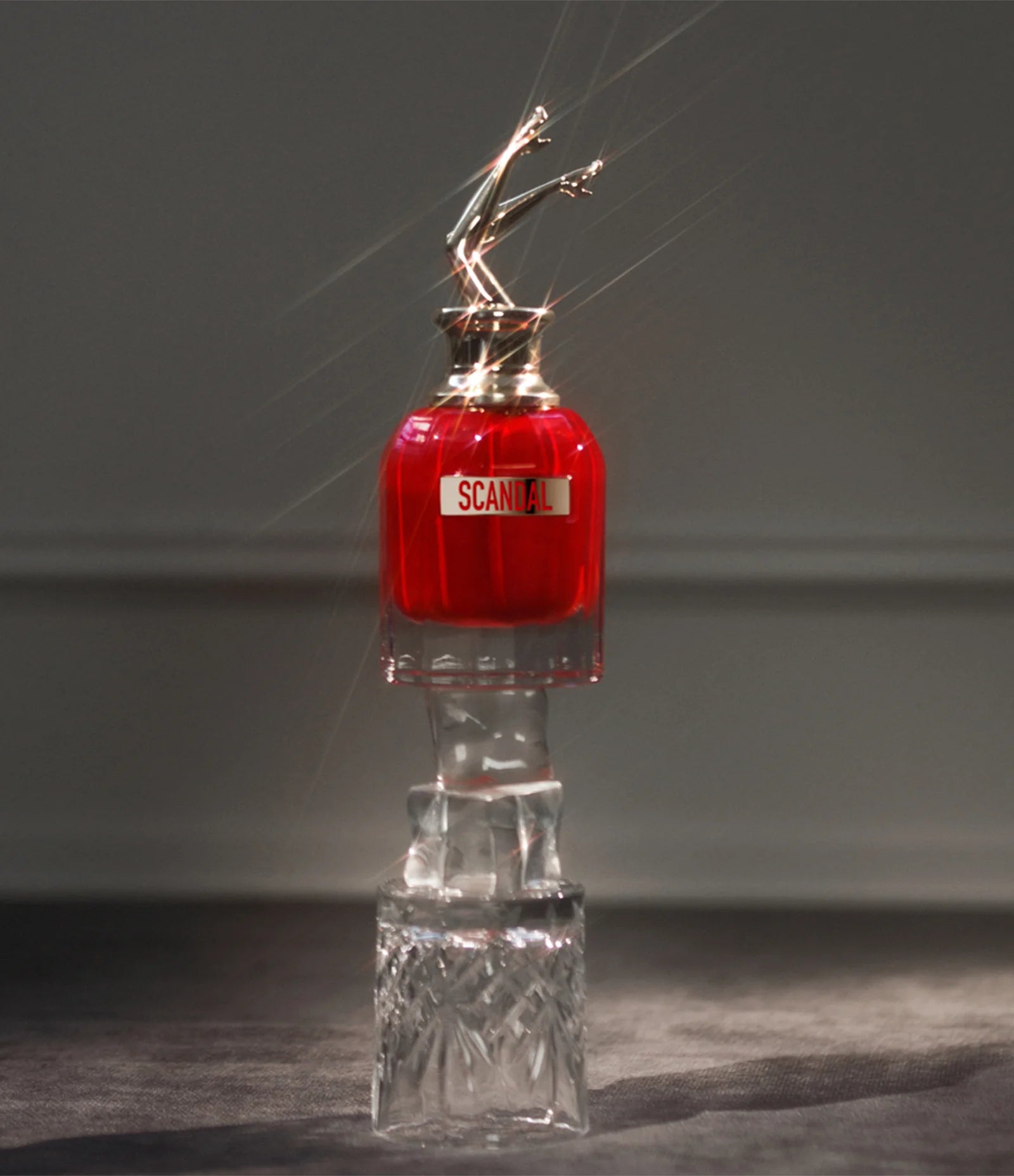 Frasco do Scandal Le Parfum iluminado por feixes de luz, posicionado sobre uma base de vidro esculpido, com destaque para o brilho intenso e sofisticado do frasco.