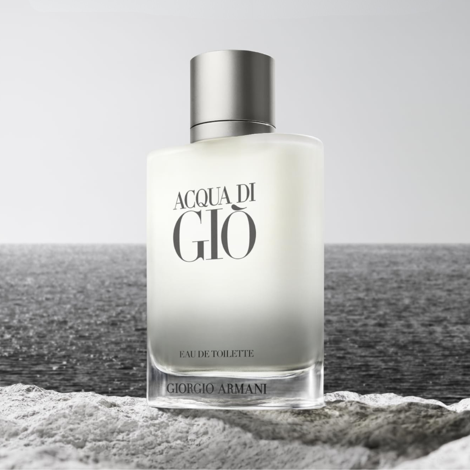 Perfume masculino Acqua di Giò Giorgio Armani em destaque sobre uma superfície de areia ou rocha clara, com o mar ou oceano ao fundo sob um céu claro, evocando uma fragrância aquática e fresca.