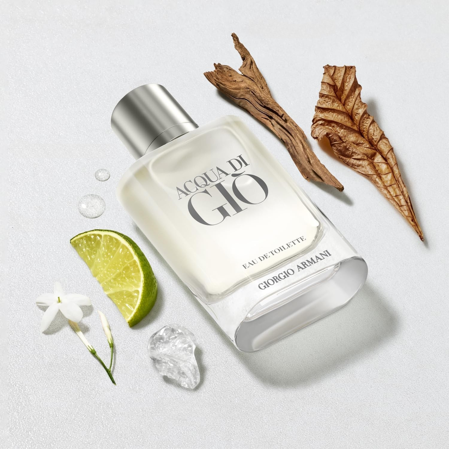 Perfume Acqua di Giò Giorgio Armani em uma composição com uma fatia de limão verde, uma flor de jasmim branca, pedaços de madeira e gotas de água, sugerindo as notas cítricas, florais e amadeiradas da fragrância, com um toque aquático.