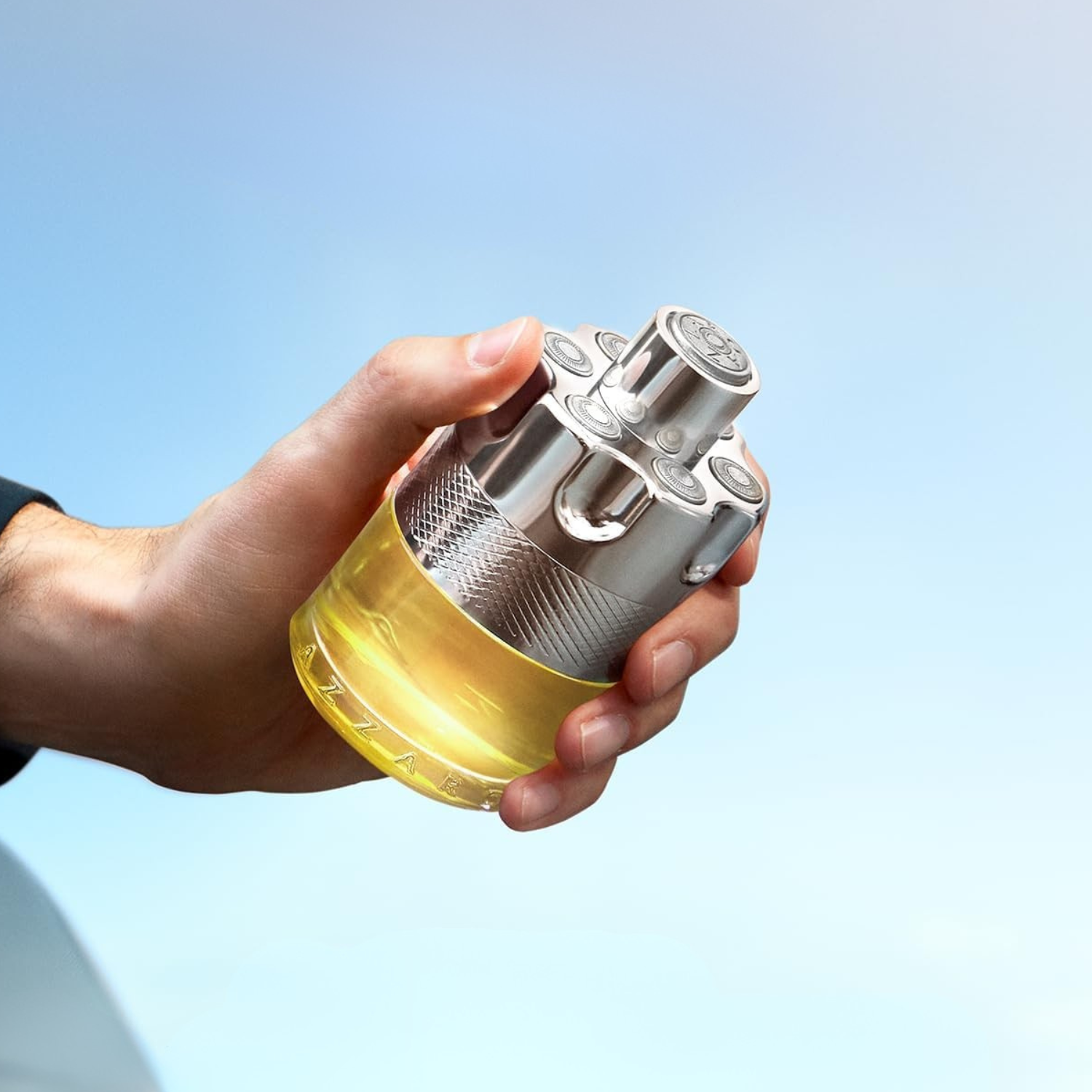Frasco do perfume Azzaro Wanted sendo segurado por uma mão masculina em frente a um céu azul. A embalagem metálica com acabamento brilhante e design robusto transmite força e masculinidade.