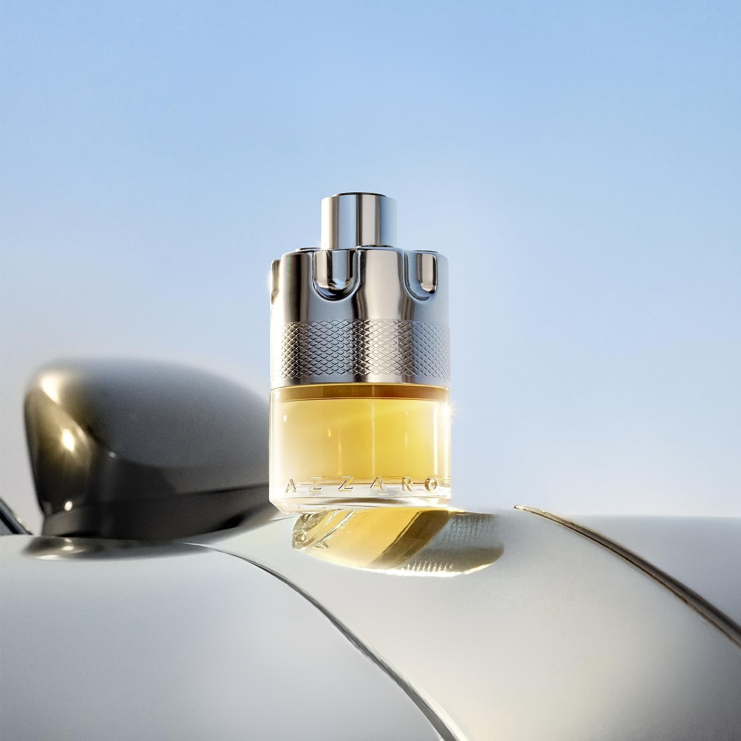 Frasco do perfume Azzaro Wanted sobre o capô de um carro prateado. O frasco reflete a luz do céu azul e reforça o estilo moderno e aventureiro da fragrância.