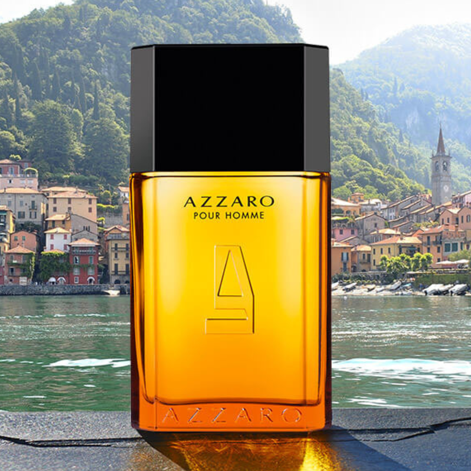 Perfume Azzaro Pour Homme posicionado à beira de um lago com vilarejo e montanhas ao fundo. O frasco âmbar reflete a luz do sol, contrastando com o cenário natural.