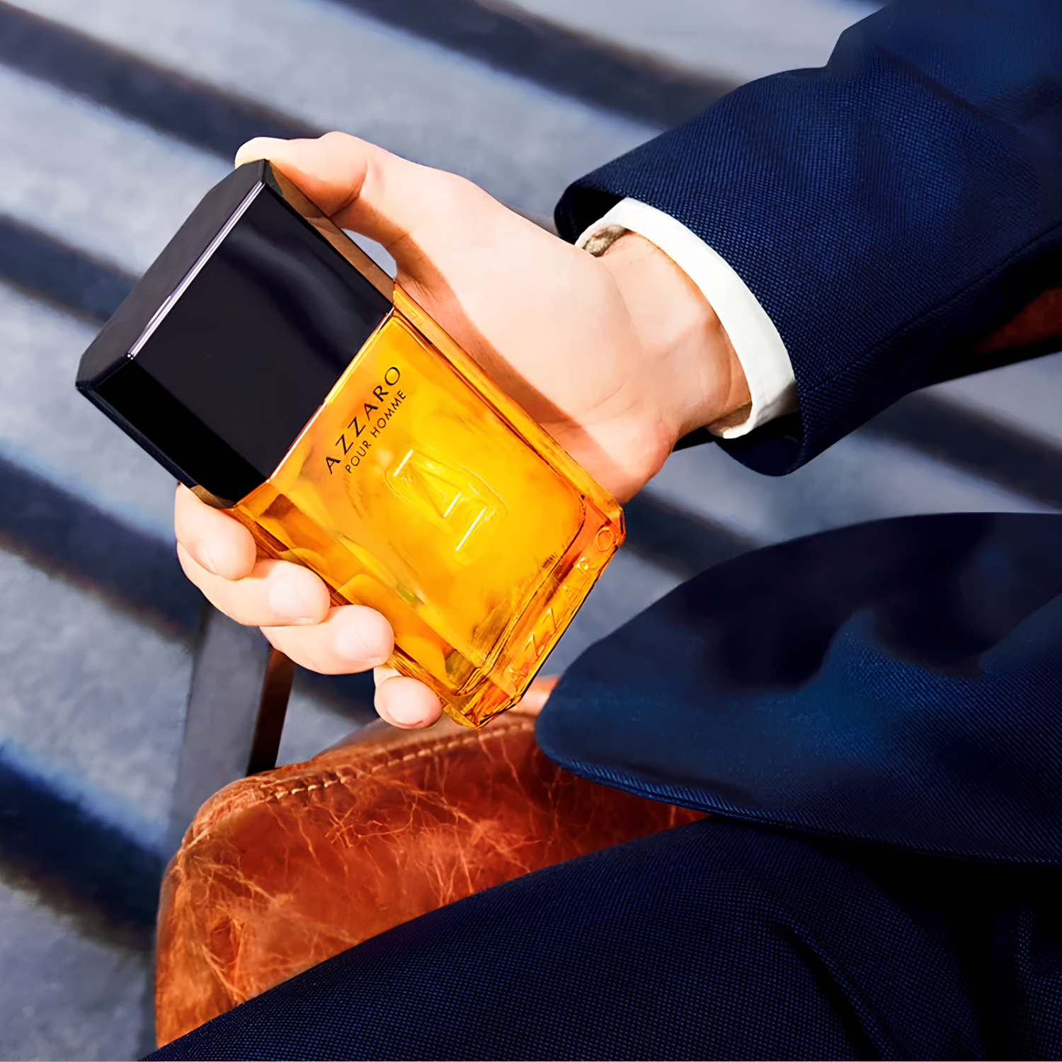 Perfume Azzaro Pour Homme sendo segurado por uma mão masculina, vestida com terno azul. O frasco âmbar e elegante está em destaque, sugerindo sofisticação e estilo.