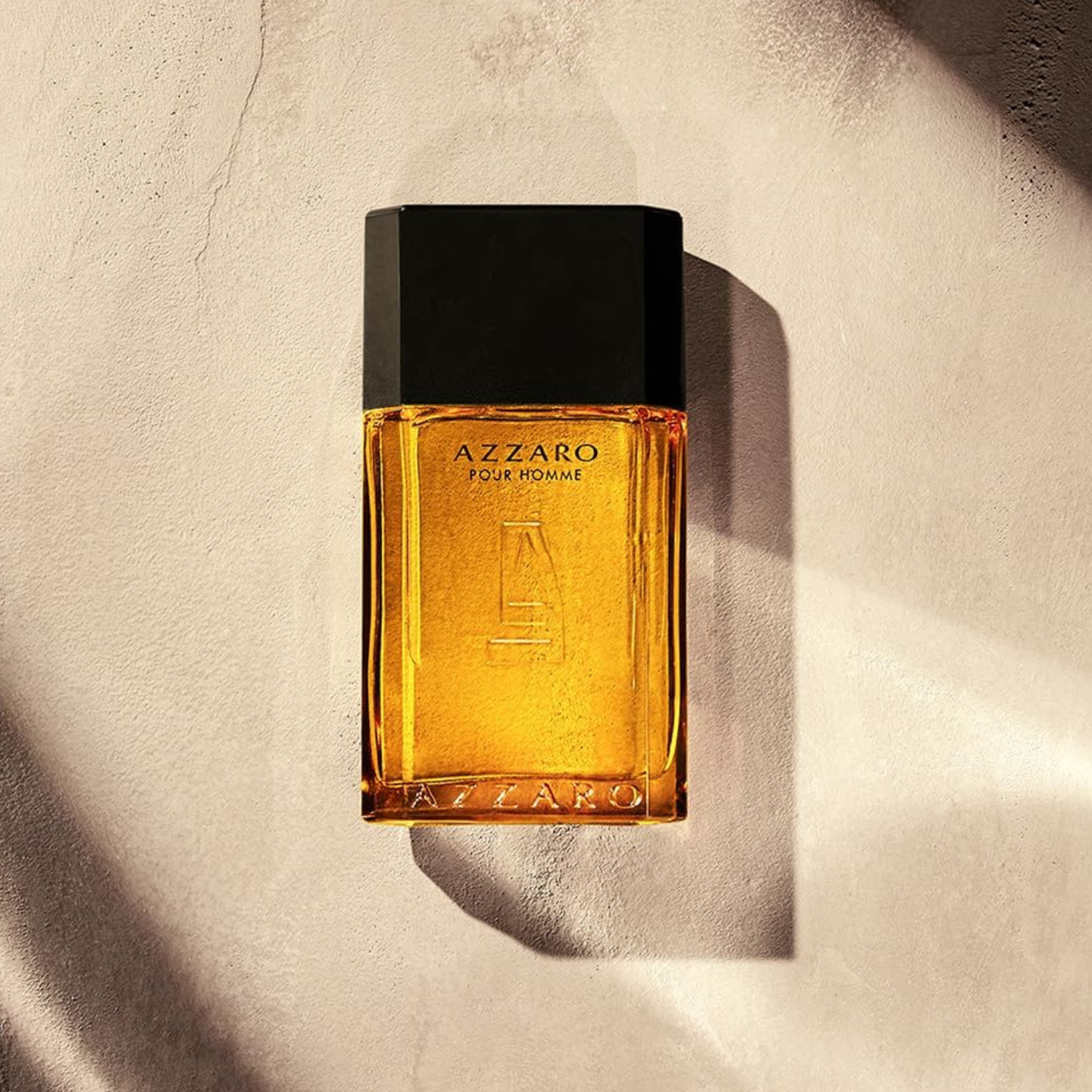 Frasco do perfume Azzaro Pour Homme deitado sobre uma superfície bege texturizada, com luz suave incidindo sobre ele. O vidro âmbar e a tampa preta contrastam com o fundo neutro.
