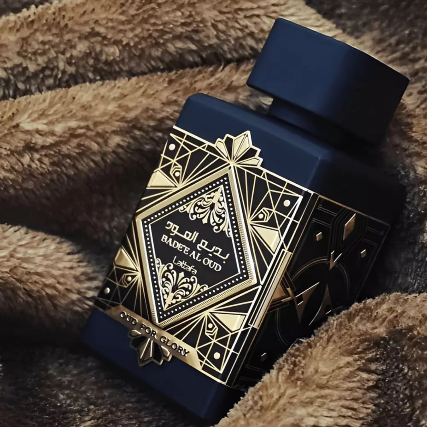 Perfume árabe Bade'e Al Oud For Glory Lattafa, à venda na Velyre, posicionado sobre tecido felpudo escuro, realçando o design dourado e a sensação de aconchego.