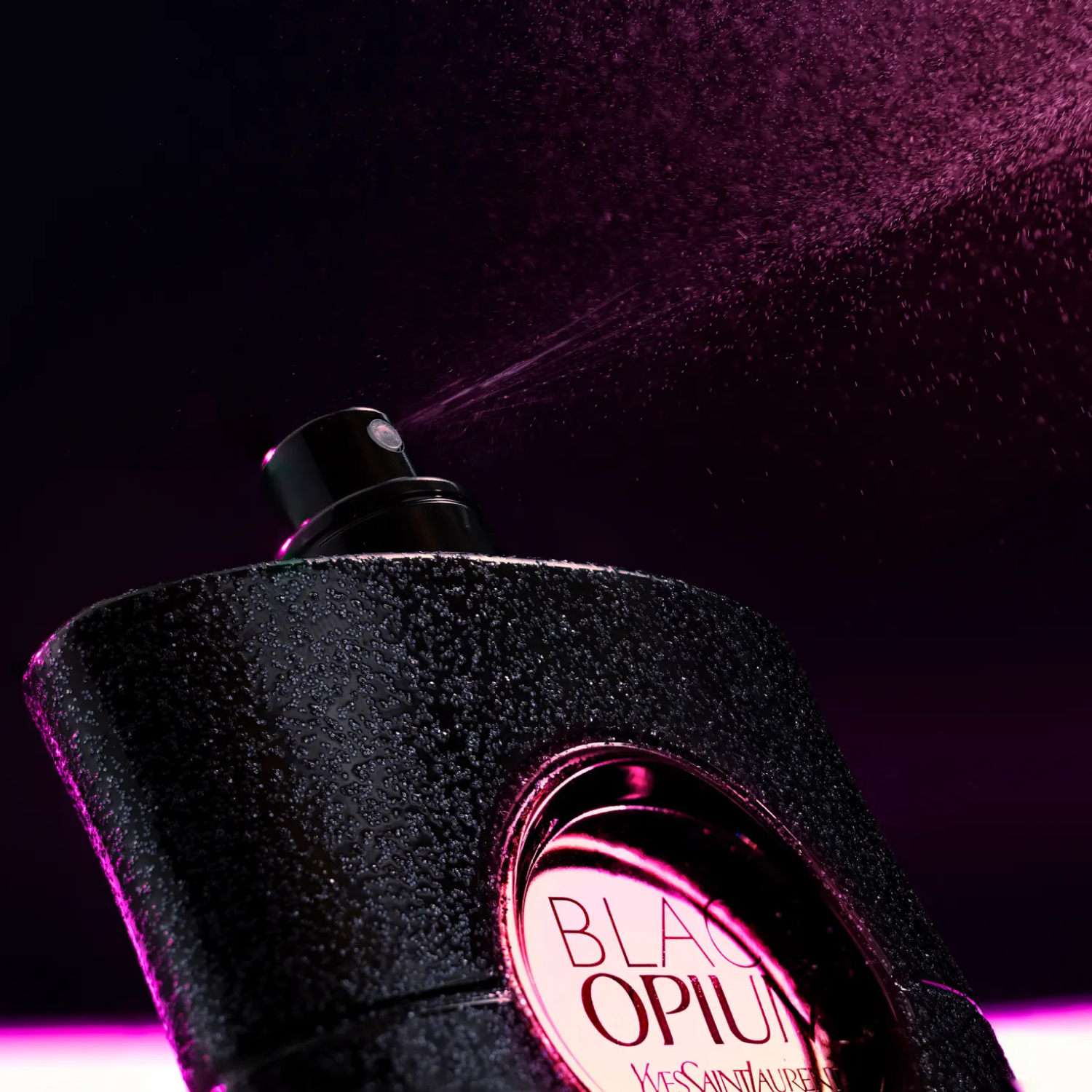 Black Opium borrifando fragrância em névoa rosa sob luz escura, com gotas visíveis sobre o frasco texturizado.