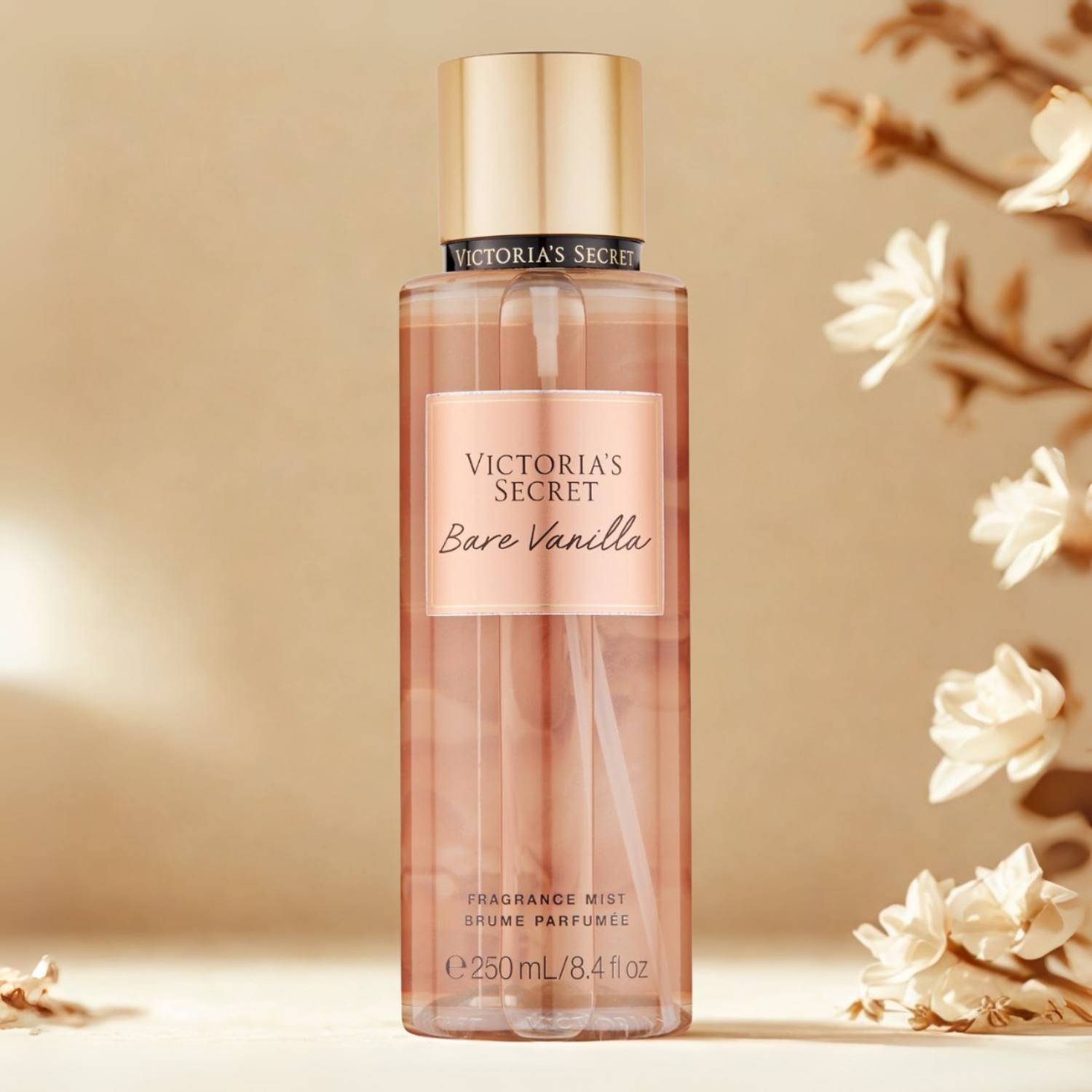 Frasco do body splash importado Bare Vanilla Victoria's Secret em destaque em um ambiente delicado com flores brancas e pétalas espalhadas, evocando uma fragrância doce e aconchegante de baunilha, disponível na Velyre.