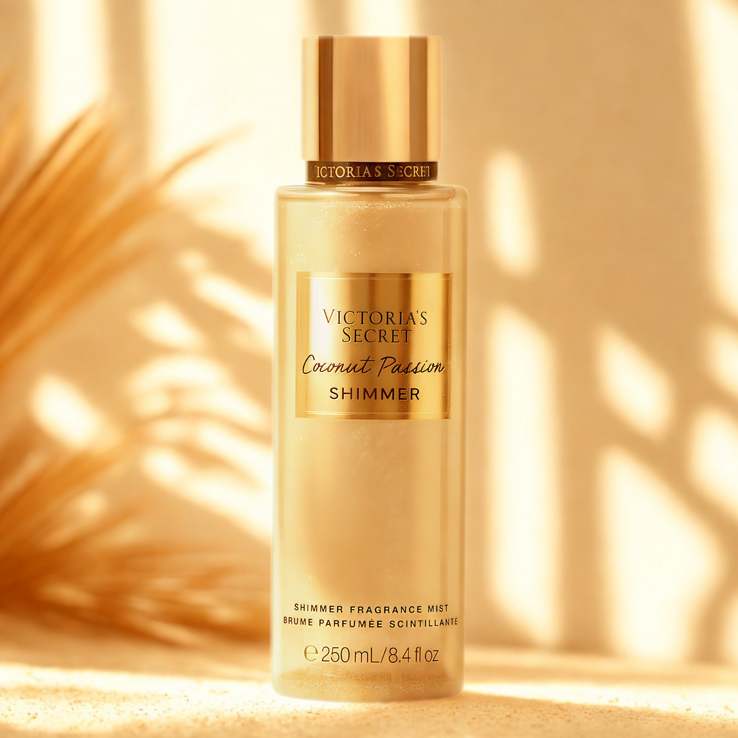 Frasco do body splash importado Coconut Passion Shimmer Victoria's Secret em destaque em um ambiente tropical com folhas de palmeira douradas ao fundo, evocando uma fragrância exótica e luminosa, disponível na Velyre.