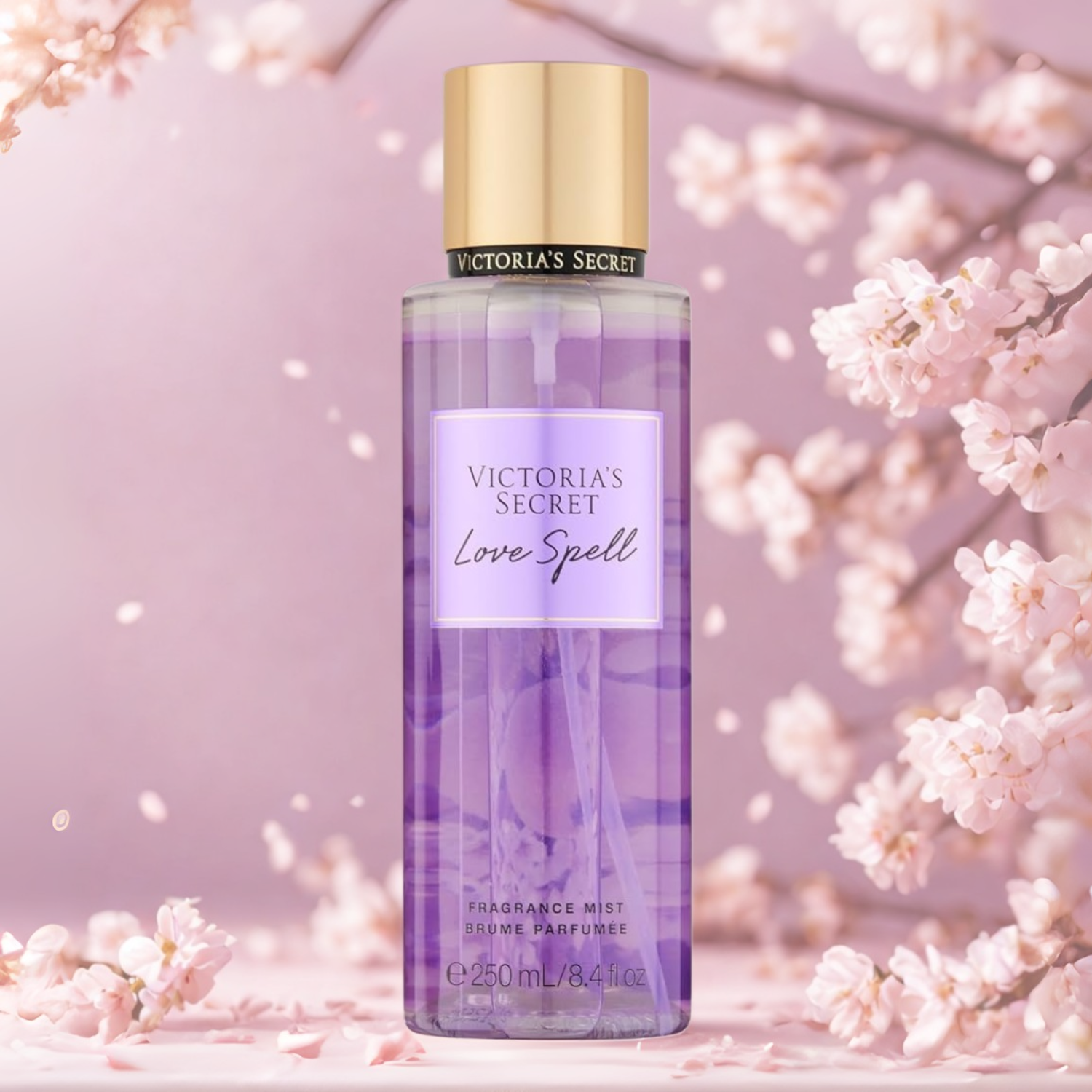 Frasco do body splash importado Love Spell Victoria's Secret em destaque em um ambiente romântico com flores de cerejeira rosa e branca ao fundo, evocando uma fragrância floral doce e romântica, disponível na Velyre.