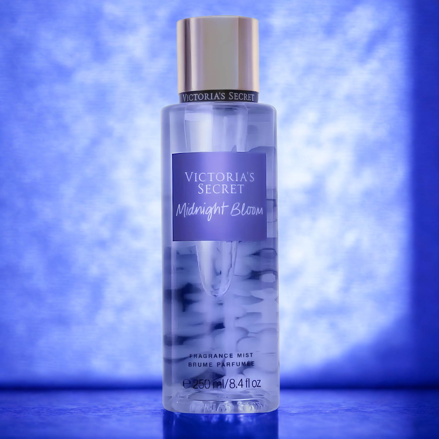 Frasco do body splash importado Midnight Bloom Victoria's Secret em destaque em um ambiente com fundo azul e luzes suaves, transmitindo uma atmosfera misteriosa e floral noturna, disponível na Velyre.