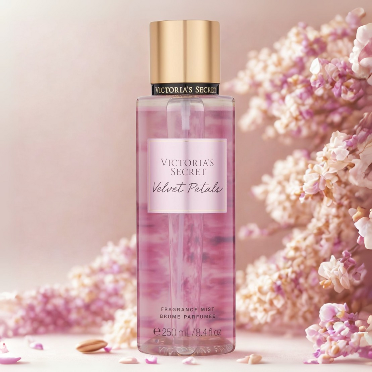 Frasco do body splash importado Velvet Petals Victoria's Secret em destaque em um ambiente romântico com volumosas flores cor-de-rosa ao fundo, evocando uma fragrância floral suave e aconchegante, disponível na Velyre.