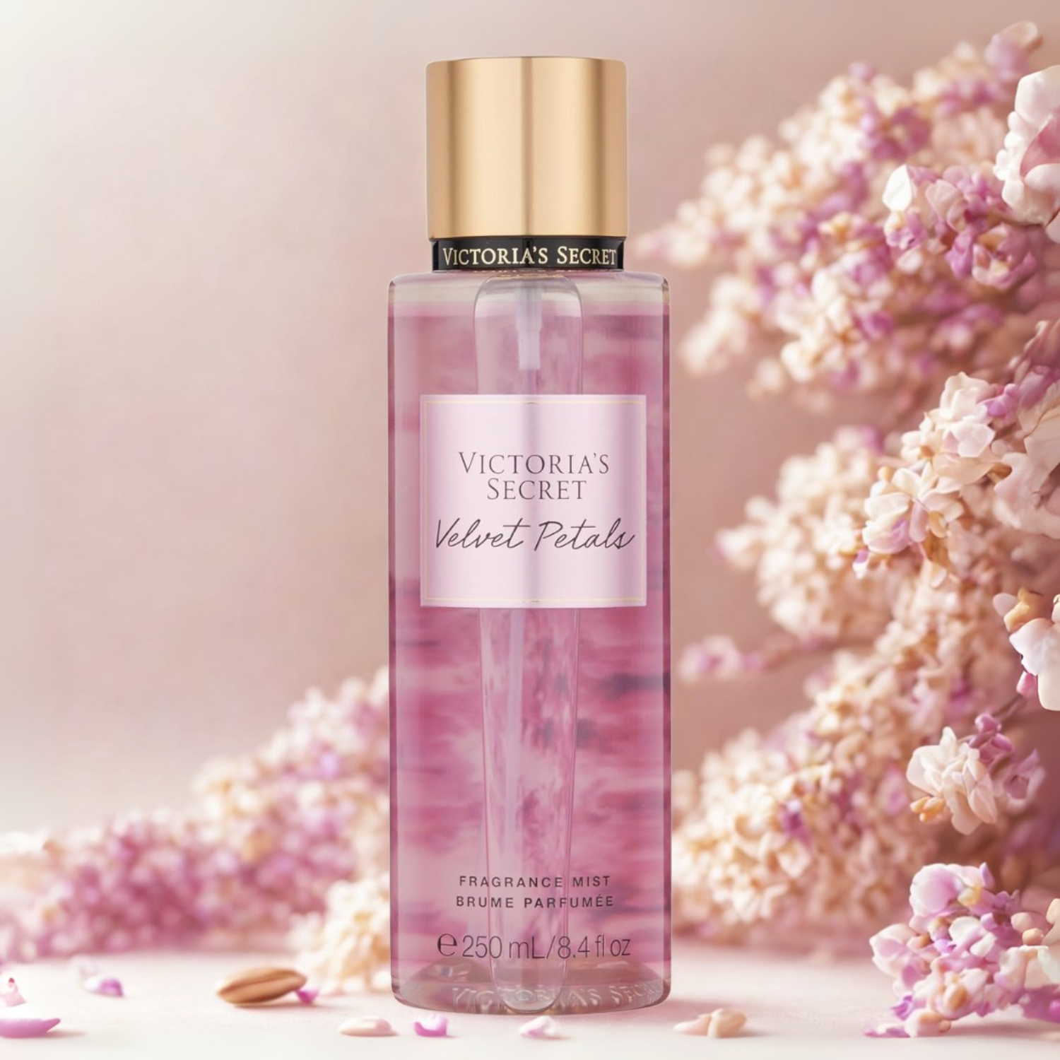 Frasco do body splash importado Velvet Petals Victoria's Secret em destaque em um ambiente romântico com volumosas flores cor-de-rosa ao fundo, evocando uma fragrância floral suave e aconchegante, disponível na Velyre.