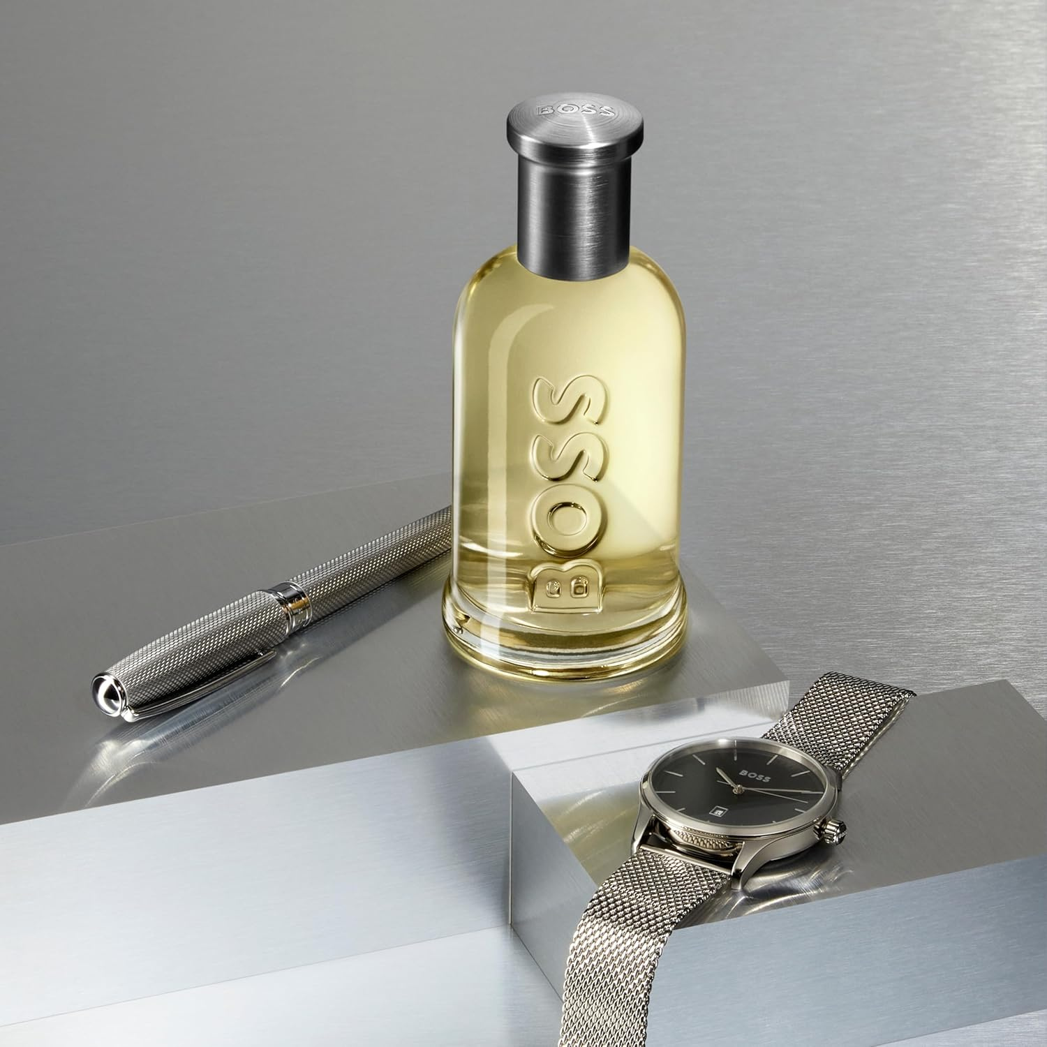 Perfume Boss Bottled posicionado em uma superfície metálica ao lado de caneta e relógio prateados, transmitindo sofisticação.