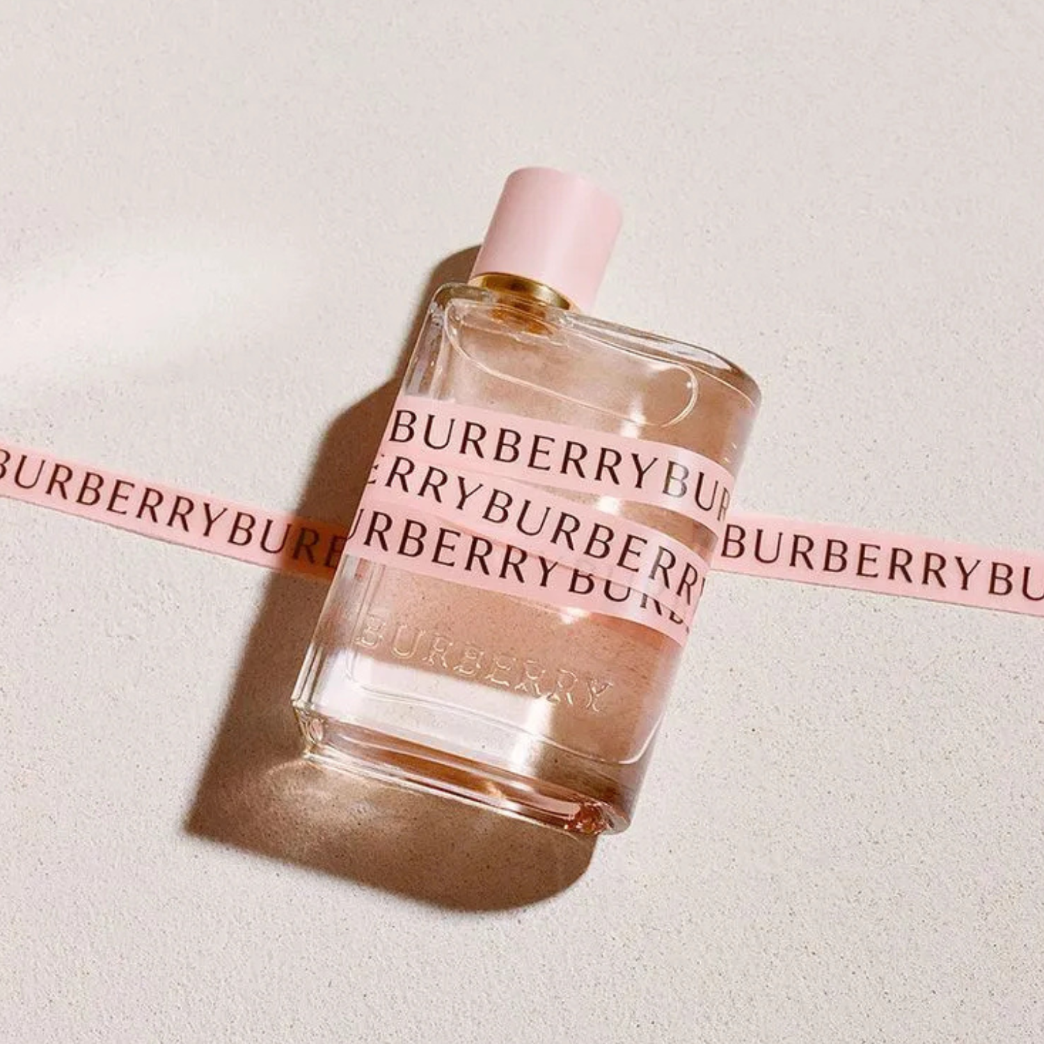 Outra vista do frasco Burberry Her sobre superfície bege com fita rosa da marca passando por trás, destacando o estilo feminino e elegante do frasco.