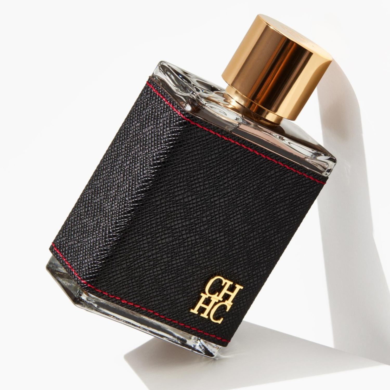 Perfume masculino CH Men Carolina Herrera inclinado sobre uma superfície branca, com uma sombra projetada, realçando o contraste do couro preto com os detalhes vermelhos e dourados, transmitindo um estilo moderno e sofisticado.
