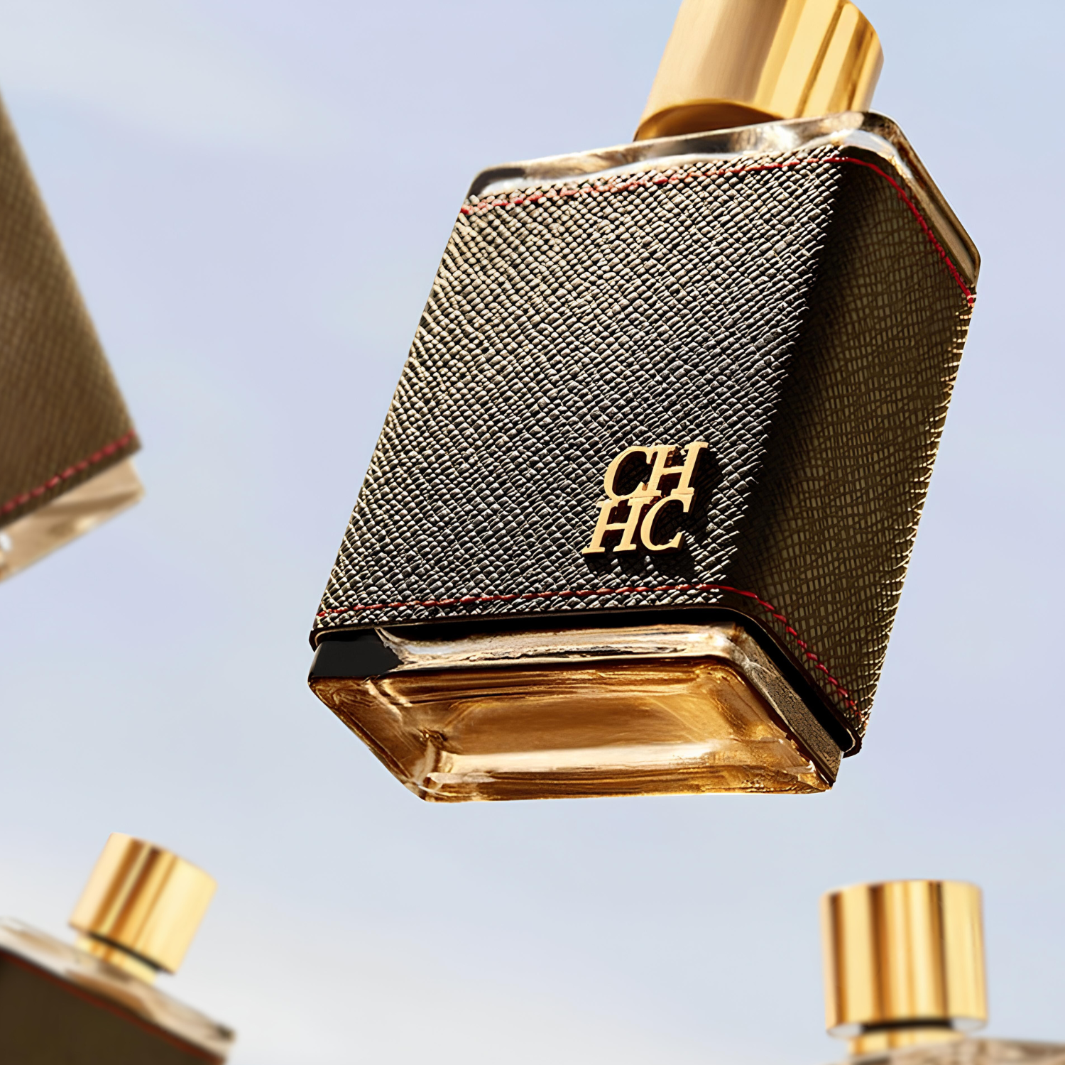 Múltiplos frascos do perfume masculino CH Men Carolina Herrera flutuando em diferentes ângulos contra um fundo azul claro, realçando o design do frasco revestido em couro preto com costuras vermelhas e o logo dourado "CH HC".