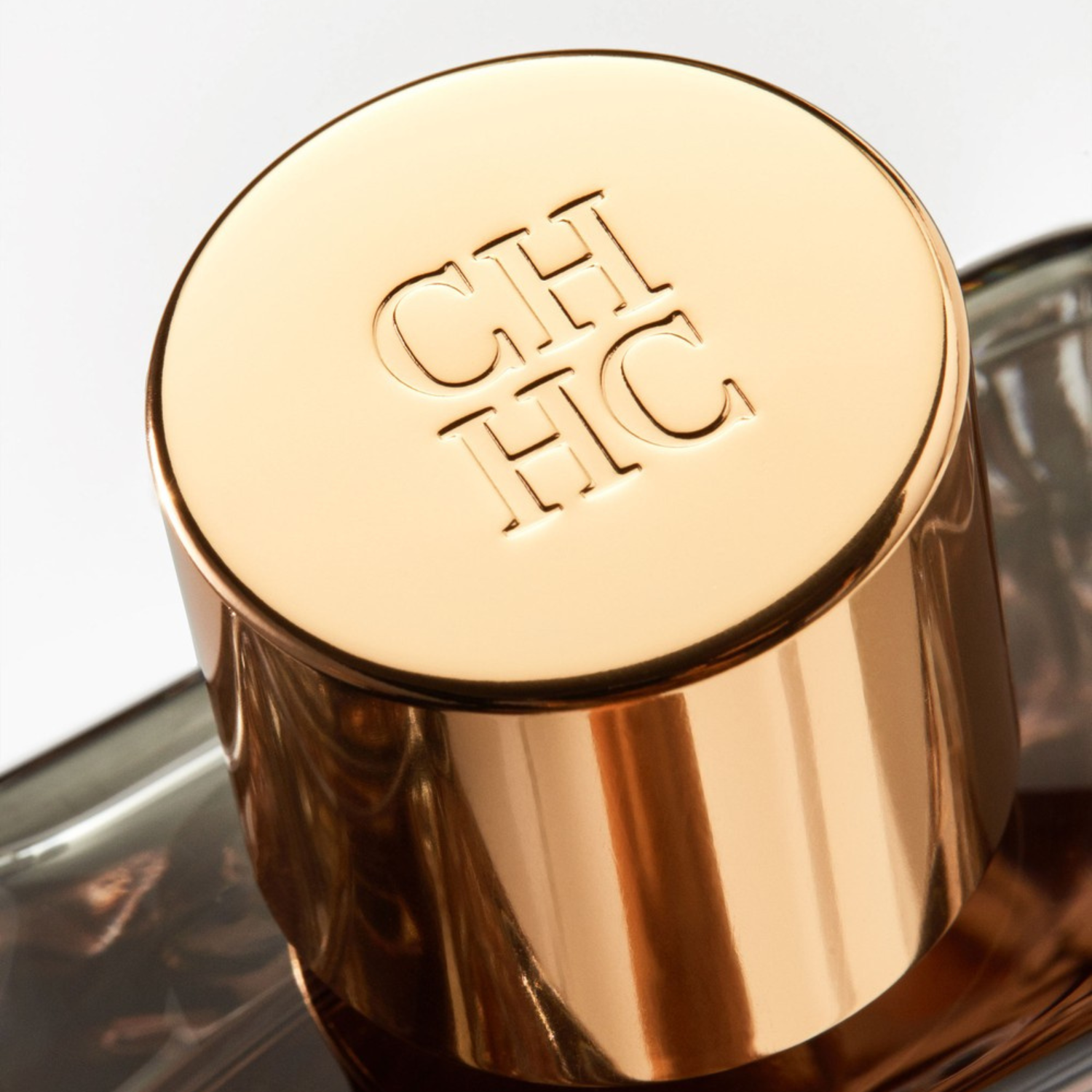 Close-up da tampa dourada e brilhante do perfume masculino CH Men Carolina Herrera, com o logo "CH HC" gravado sutilmente na superfície, demonstrando o detalhe e a elegância da embalagem.