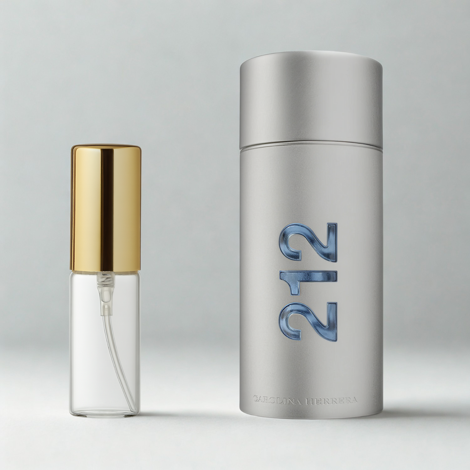 Decant de 5ml com borrifador dourado ao lado do frasco original do perfume importado masculino 212 Men Carolina Herrera Eau de Toilette, ambos sobre uma superfície e fundo cinza claro. A imagem destaca a apresentação do decant para venda e a familiaridade com o frasco original, disponíveis na Velyre.