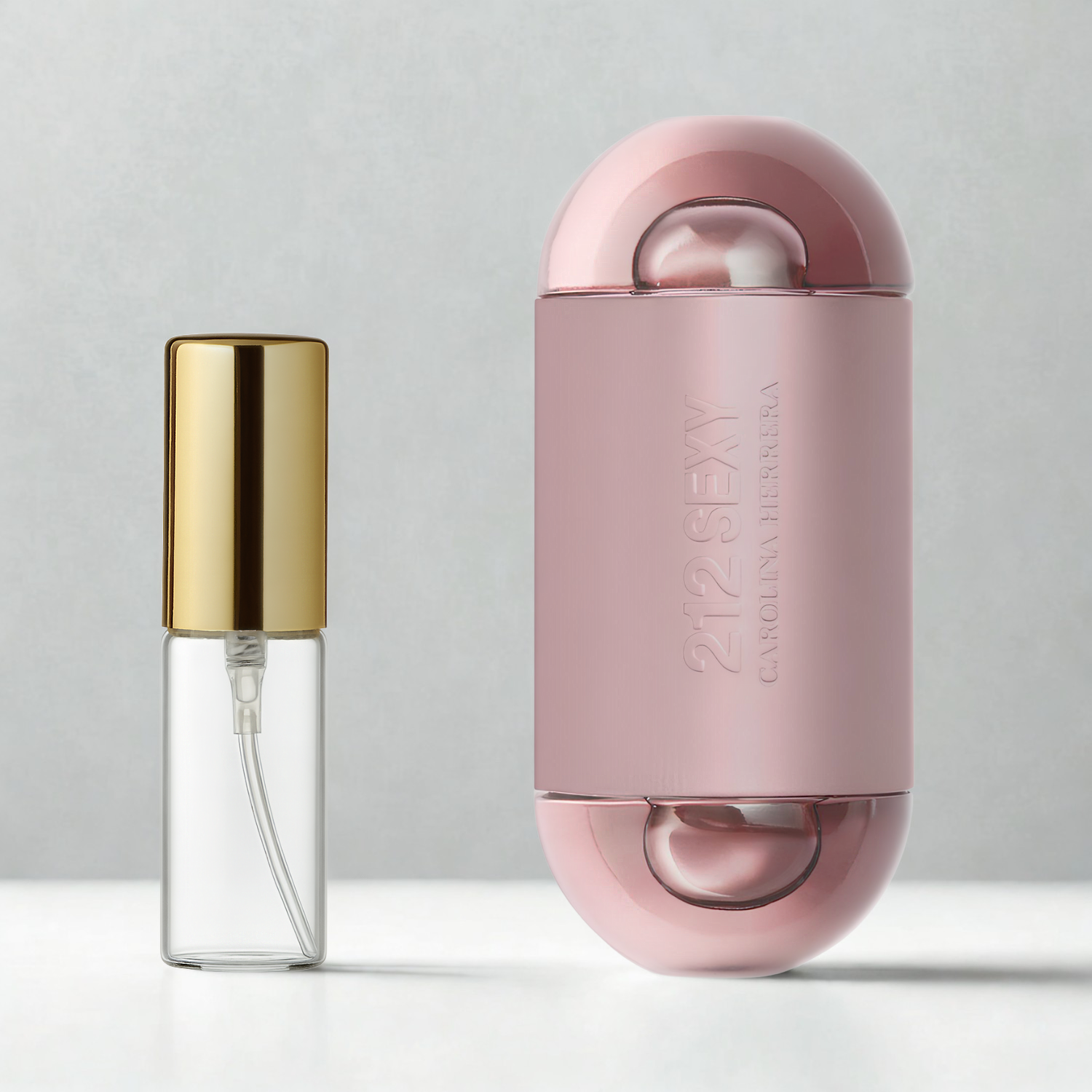 Decant de 5ml com borrifador dourado ao lado do frasco original do perfume importado feminino 212 Sexy Carolina Herrera Eau de Parfum, ambos sobre uma superfície e fundo cinza claro. A imagem destaca a apresentação do decant para venda e a familiaridade com o frasco original, disponíveis na Velyre.