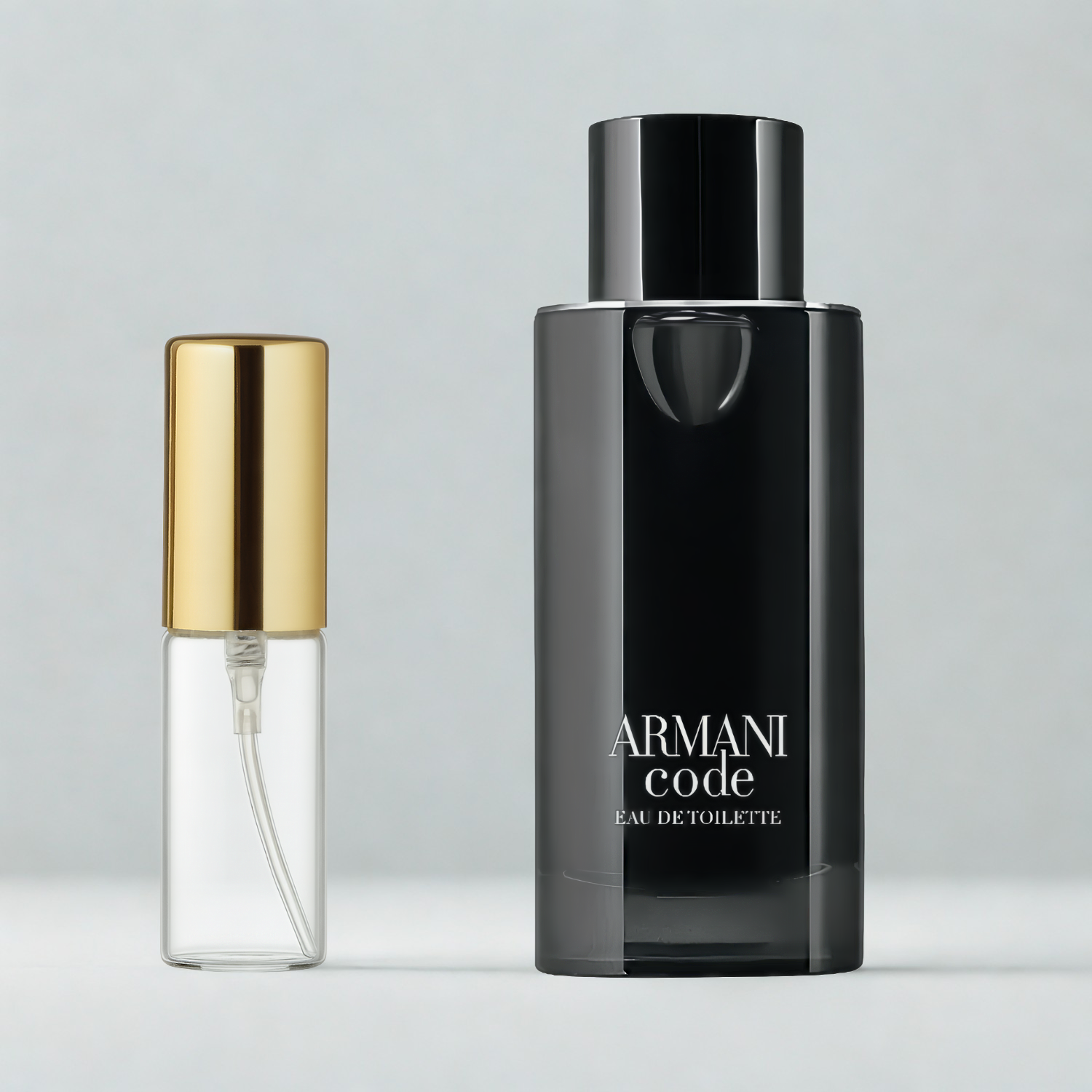 Decant de 5ml com borrifador dourado ao lado do frasco original do perfume importado masculino Armani Code Giorgio Armani Eau de Toilette, ambos sobre uma superfície e fundo cinza claro. A imagem destaca a apresentação do decant para venda e a familiaridade com o frasco original, disponíveis na Velyre.