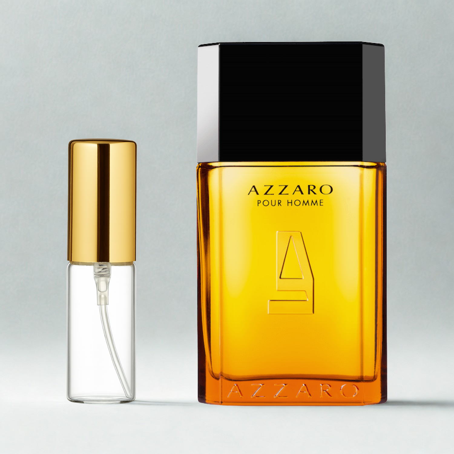 Decant de 5ml com borrifador dourado ao lado do frasco original do perfume importado masculino Azzaro Pour Homme Eau de Toilette, ambos sobre uma superfície e fundo cinza claro. A imagem destaca a apresentação do decant para venda e a familiaridade com o frasco original, disponíveis na Velyre.