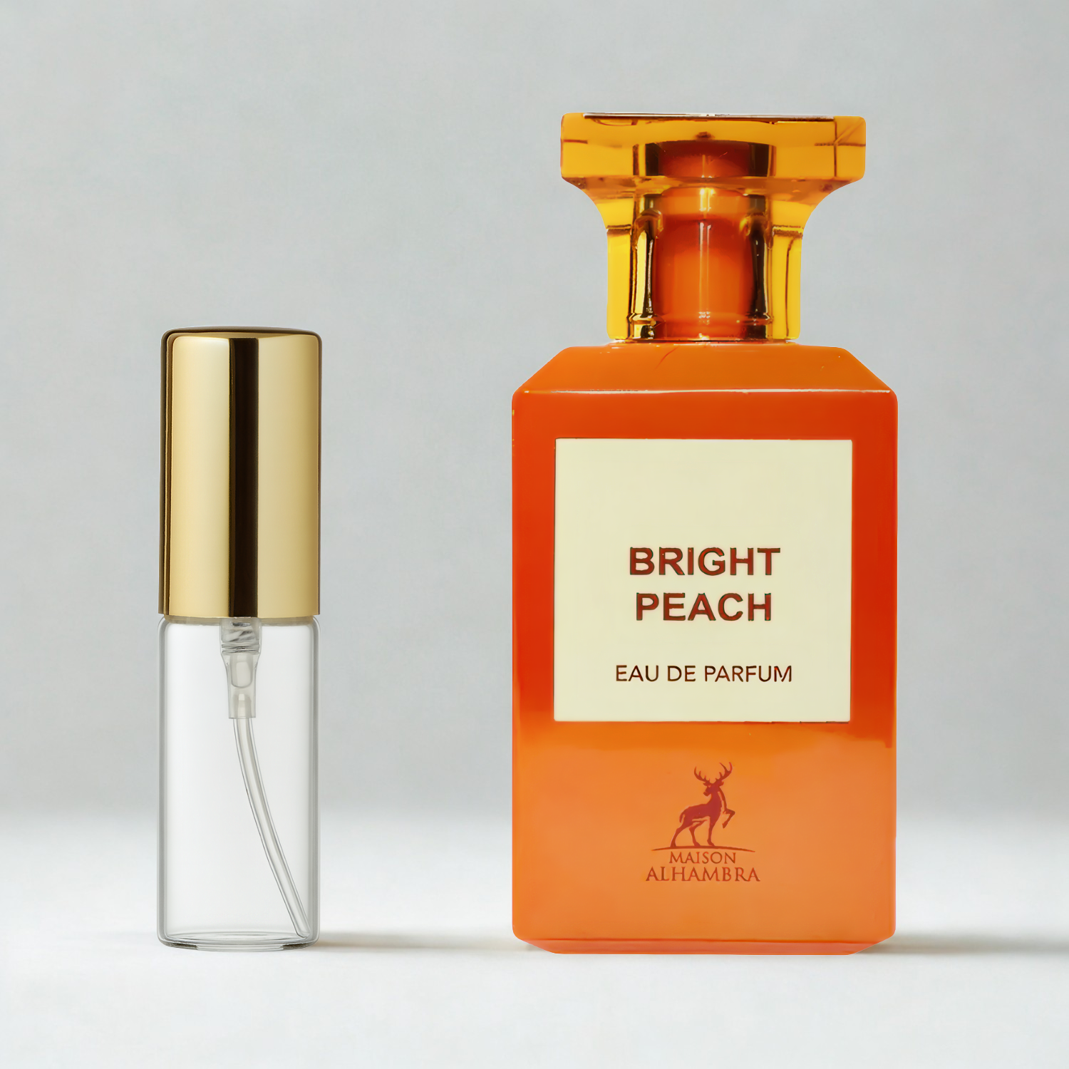 Decant de 5ml com borrifador dourado ao lado do frasco original do perfume árabe unissex Bright Peach Maison Alhambra Eau de Parfum, ambos sobre uma superfície e fundo cinza claro. A imagem destaca a apresentação do decant para venda e a familiaridade com o frasco original, disponíveis na Velyre.