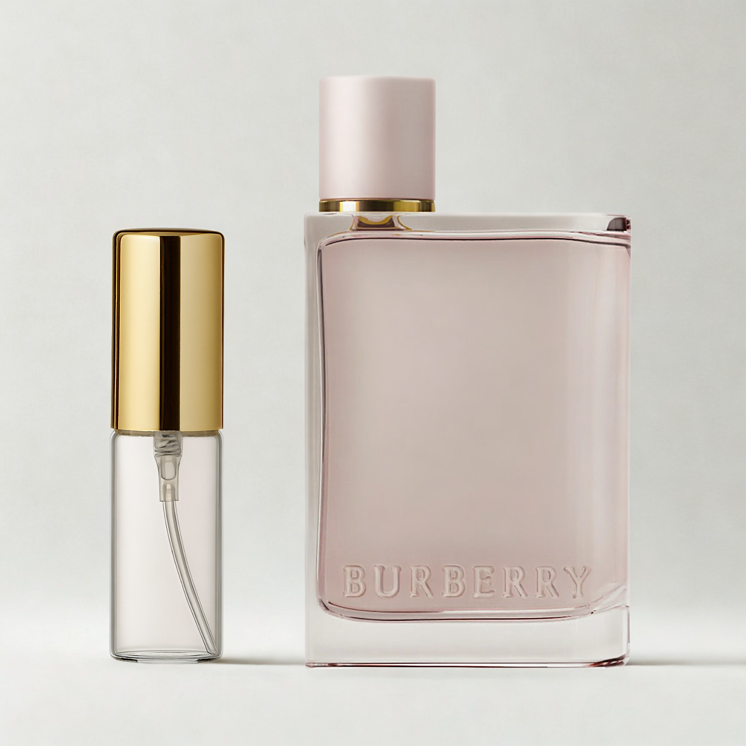Decant de 5ml com borrifador dourado ao lado do frasco original do perfume importado feminino Burberry Her Eau de Parfum, ambos sobre uma superfície e fundo cinza claro. A imagem destaca a apresentação do decant para venda e a familiaridade com o frasco original, disponíveis na Velyre.