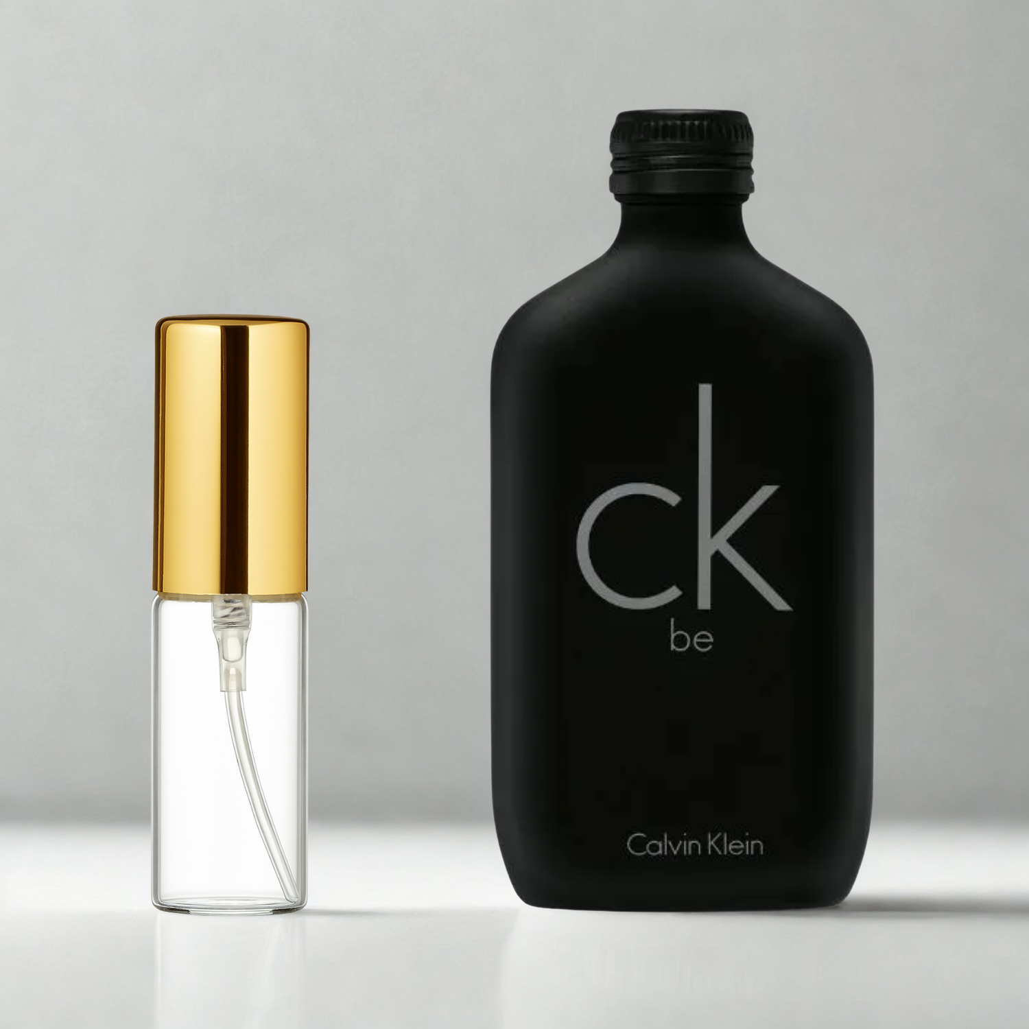 Decant de 5ml com borrifador dourado ao lado do frasco original do perfume importado unissex CK Be Calvin Klein Eau de Toilette, ambos sobre uma superfície e fundo cinza claro. A imagem destaca a apresentação do decant para venda e a familiaridade com o frasco original, disponíveis na Velyre.