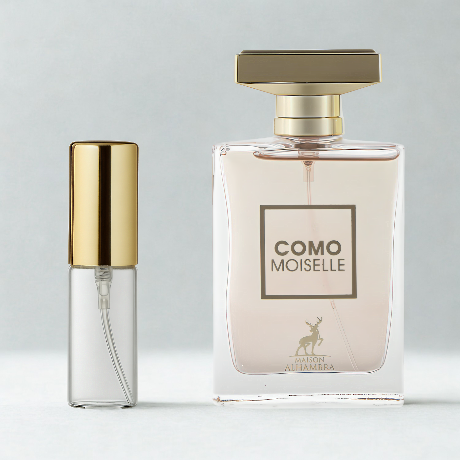 Decant de 5ml com borrifador dourado ao lado do frasco original do perfume árabe feminino Como Moiselle Maison Alhambra Eau de Parfum, ambos sobre uma superfície e fundo cinza claro. A imagem destaca a apresentação do decant para venda e a familiaridade com o frasco original, disponíveis na Velyre.