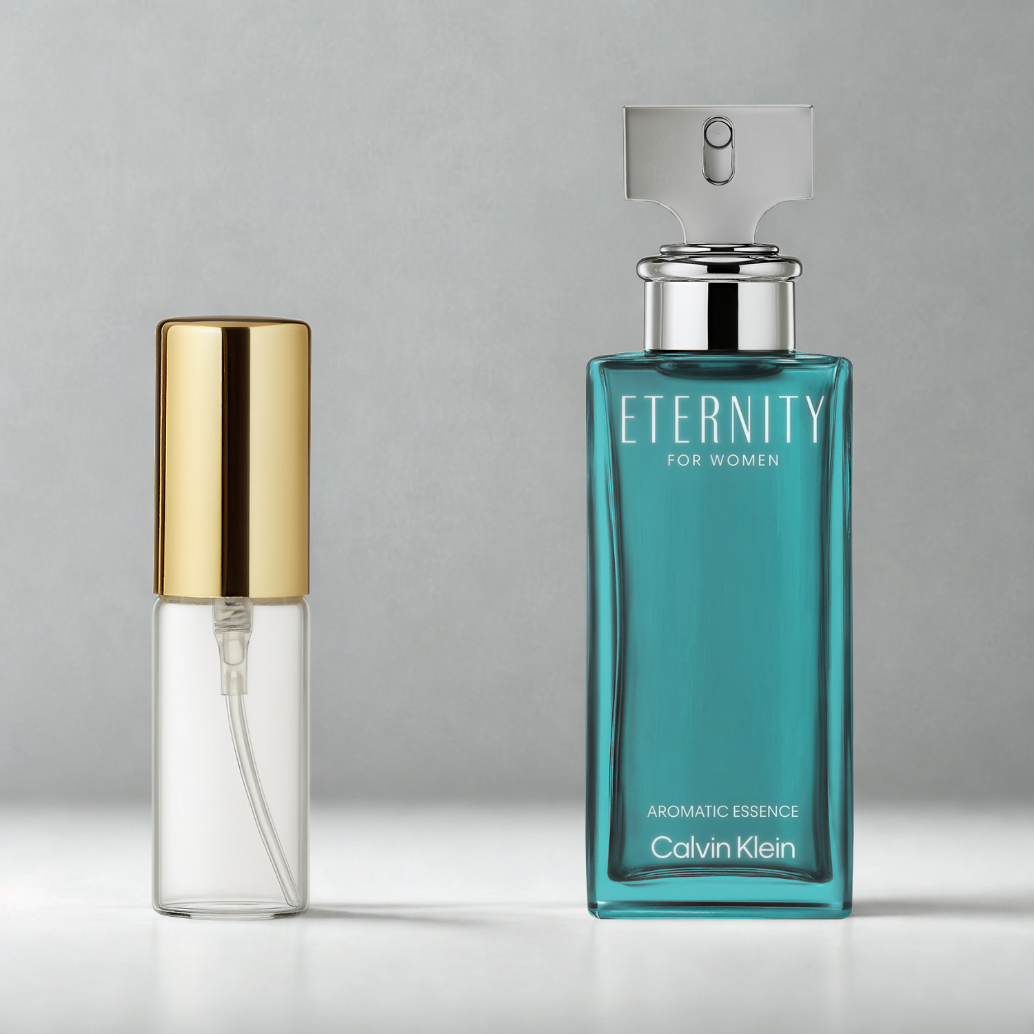 Decant de 5ml com borrifador dourado ao lado do frasco original do perfume importado feminino Eternity Aromatic Essence for Women Calvin Klein Eau de Parfum, ambos sobre uma superfície e fundo cinza claro. A imagem destaca a apresentação do decant para venda e a familiaridade com o frasco original, disponíveis na Velyre.