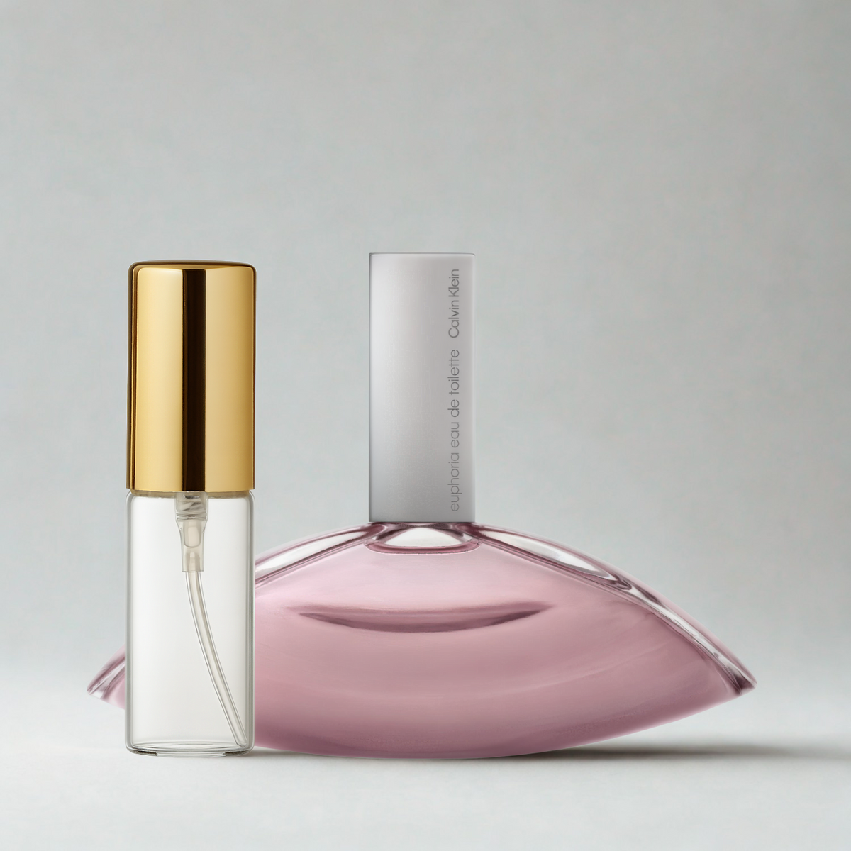 Decant de 5ml com borrifador dourado ao lado do frasco original do perfume importado feminino Euphoria for Women Calvin Klein Eau de Toilette, ambos sobre uma superfície e fundo cinza claro. A imagem destaca a apresentação do decant para venda e a familiaridade com o frasco original, disponíveis na Velyre.