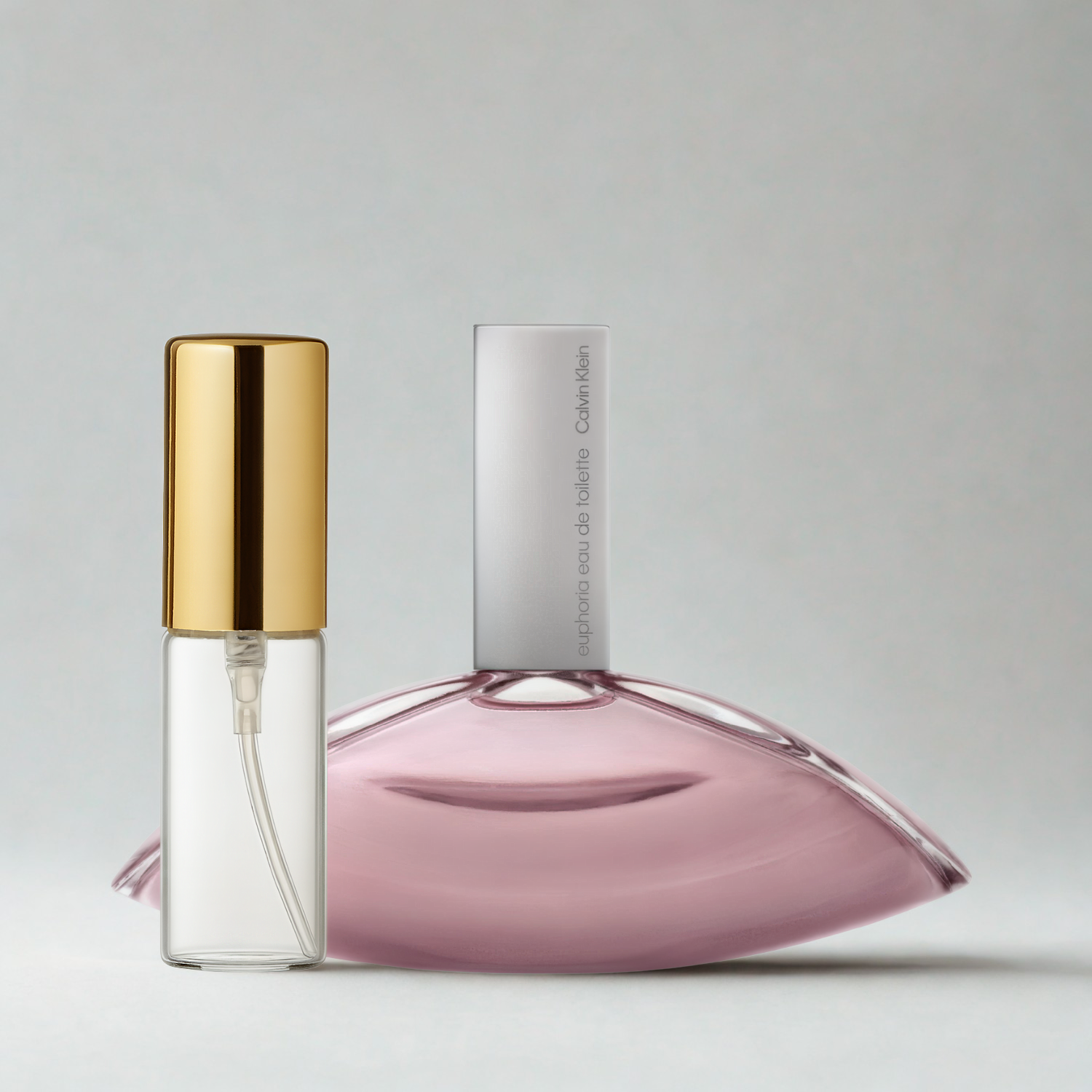 Decant de 5ml com borrifador dourado ao lado do frasco original do perfume importado feminino Euphoria for Women Calvin Klein Eau de Toilette, ambos sobre uma superfície e fundo cinza claro. A imagem destaca a apresentação do decant para venda e a familiaridade com o frasco original, disponíveis na Velyre.