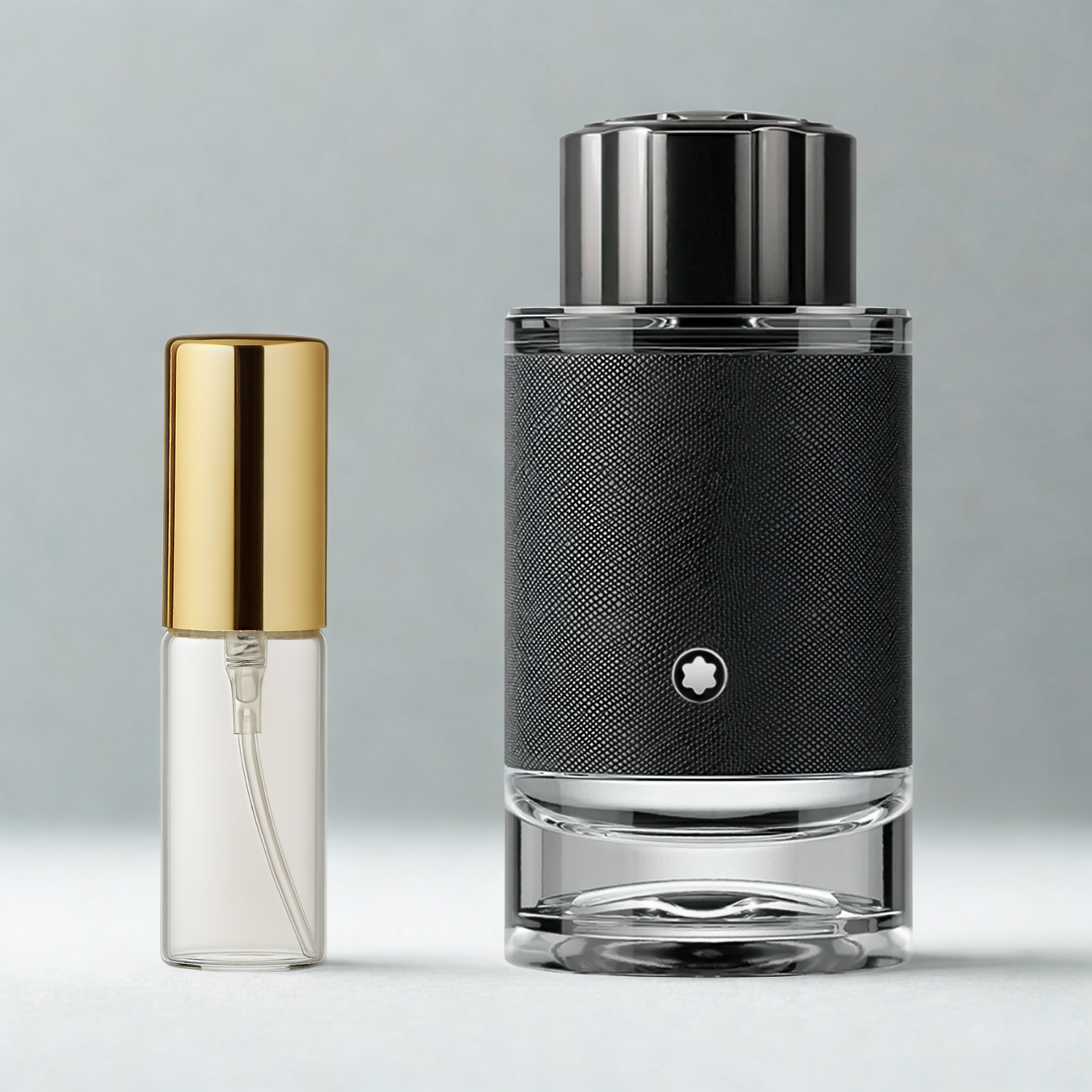 Decant de 5ml com borrifador dourado ao lado do frasco original do perfume importado masculino Explorer Montblanc Eau de Parfum, ambos sobre uma superfície e fundo cinza claro. A imagem destaca a apresentação do decant para venda e a familiaridade com o frasco original, disponíveis na Velyre.