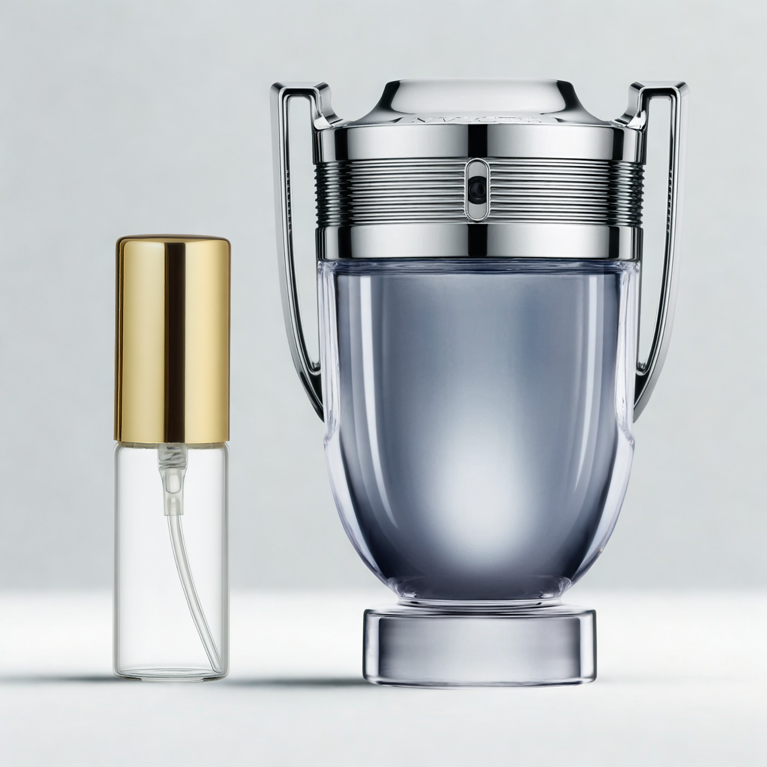 Decant de 5ml com borrifador dourado ao lado do frasco original do perfume importado masculino Invictus Rabanne Eau de Toilette, ambos sobre uma superfície e fundo cinza claro. A imagem destaca a apresentação do decant para venda e a familiaridade com o frasco original, disponíveis na Velyre.