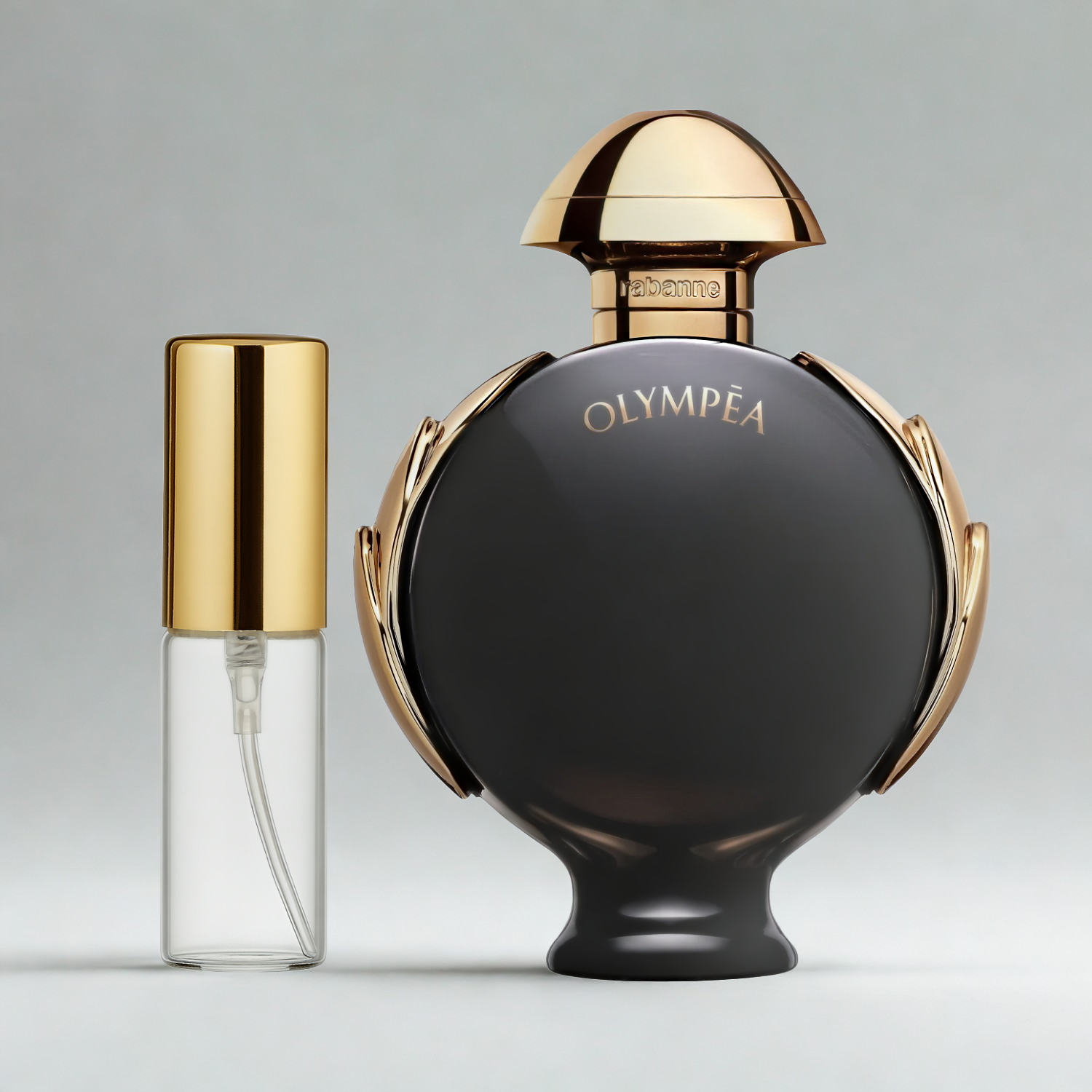 Decant de 5ml com borrifador dourado ao lado do frasco original do perfume importado feminino Olympéa Parfum Rabanne Eau de Parfum, ambos sobre uma superfície e fundo cinza claro. A imagem destaca a apresentação do decant para venda e a familiaridade com o frasco original, disponíveis na Velyre.