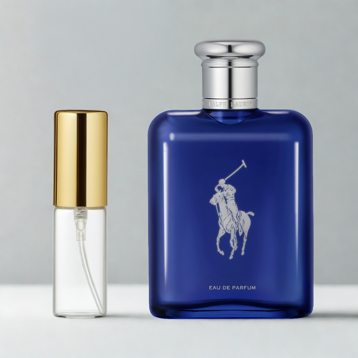 Decant de 5ml com borrifador dourado ao lado do frasco original do perfume importado masculino Polo Blue Ralph Lauren Eau de Parfum, ambos sobre uma superfície e fundo cinza claro. A imagem destaca a apresentação do decant para venda e a familiaridade com o frasco original, disponíveis na Velyre.