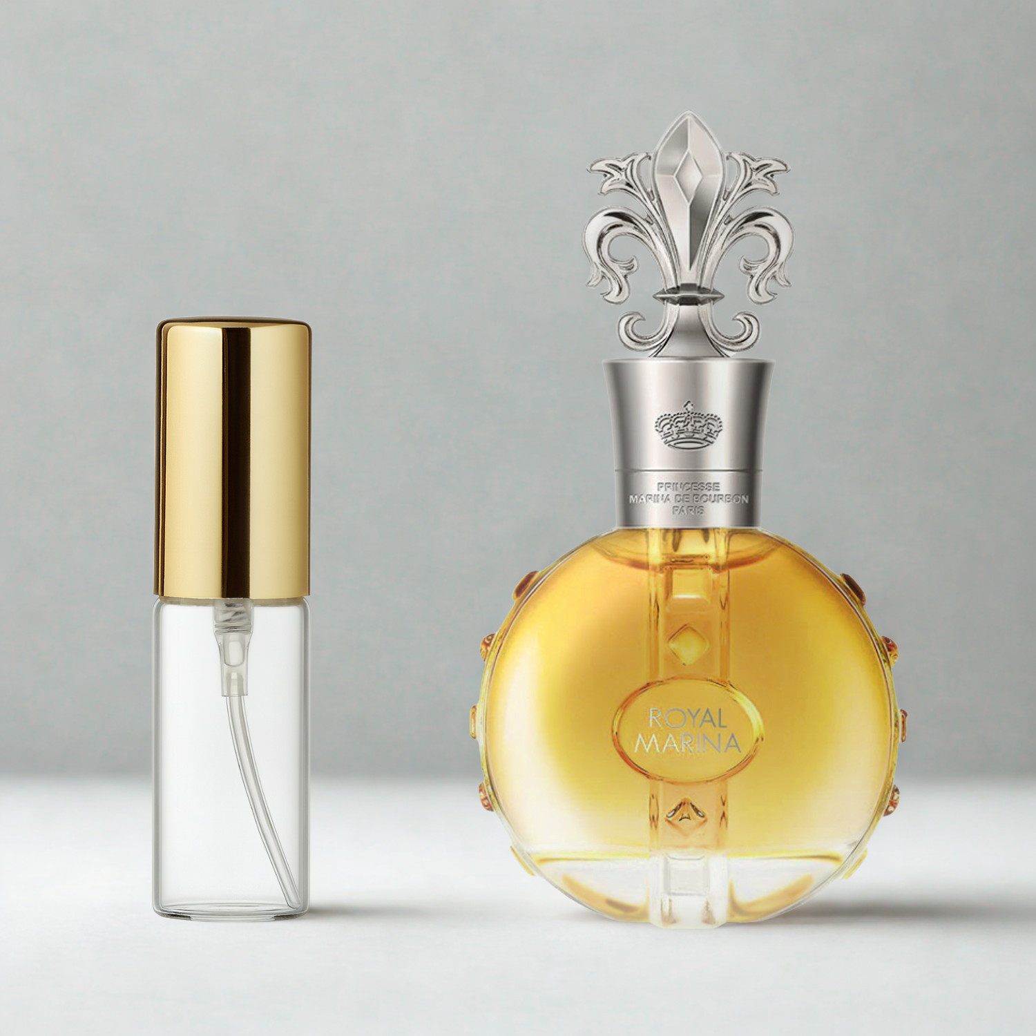 Decant de 5ml com borrifador dourado ao lado do frasco original do perfume importado feminino Royal Marina Diamond Marina de Bourbon Eau de Parfum, ambos sobre uma superfície e fundo cinza claro. A imagem destaca a apresentação do decant para venda e a familiaridade com o frasco original, disponíveis na Velyre.