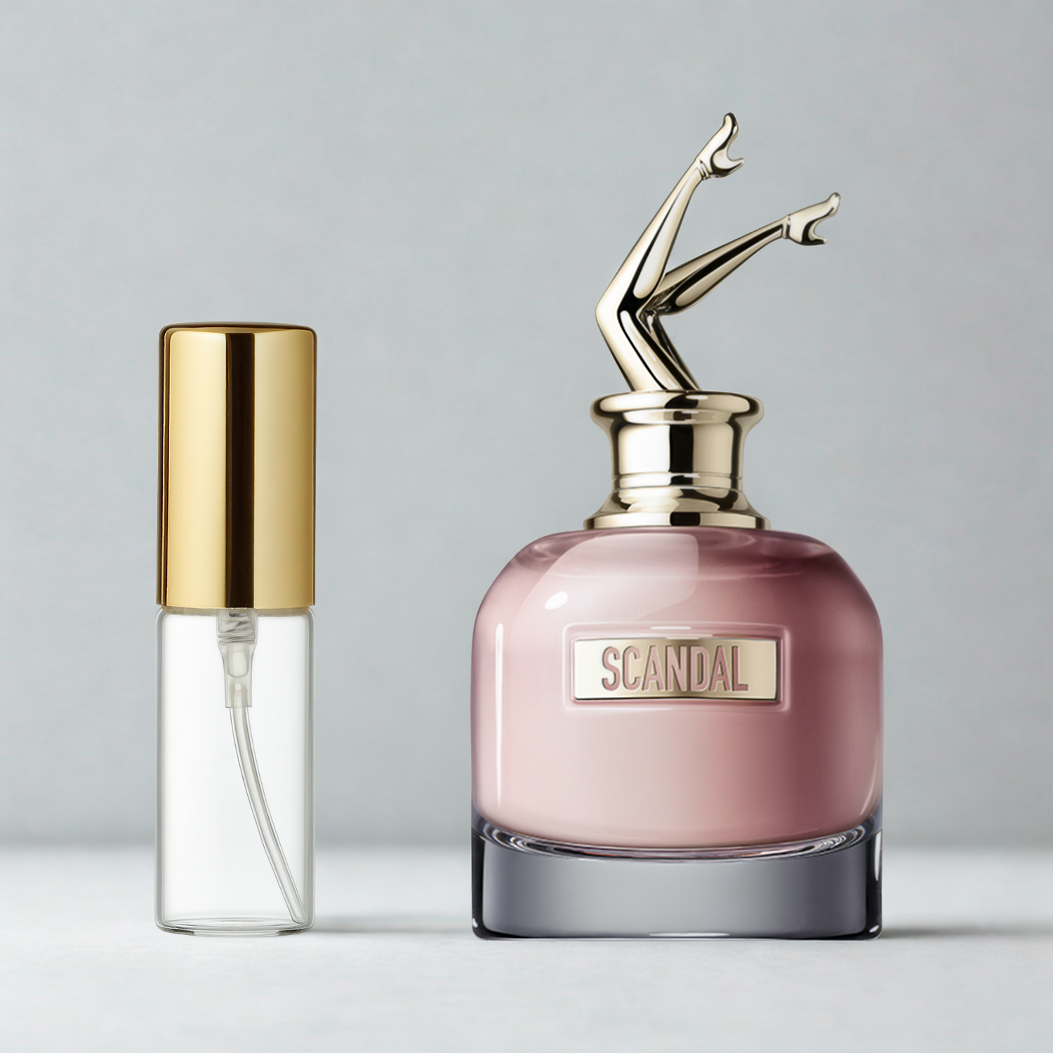 Decant de 5ml com borrifador dourado ao lado do frasco original do perfume importado feminino Scandal Jean Paul Gaultier Eau de Parfum, ambos sobre uma superfície e fundo cinza claro. A imagem destaca a apresentação do decant para venda e a familiaridade com o frasco original, disponíveis na Velyre.